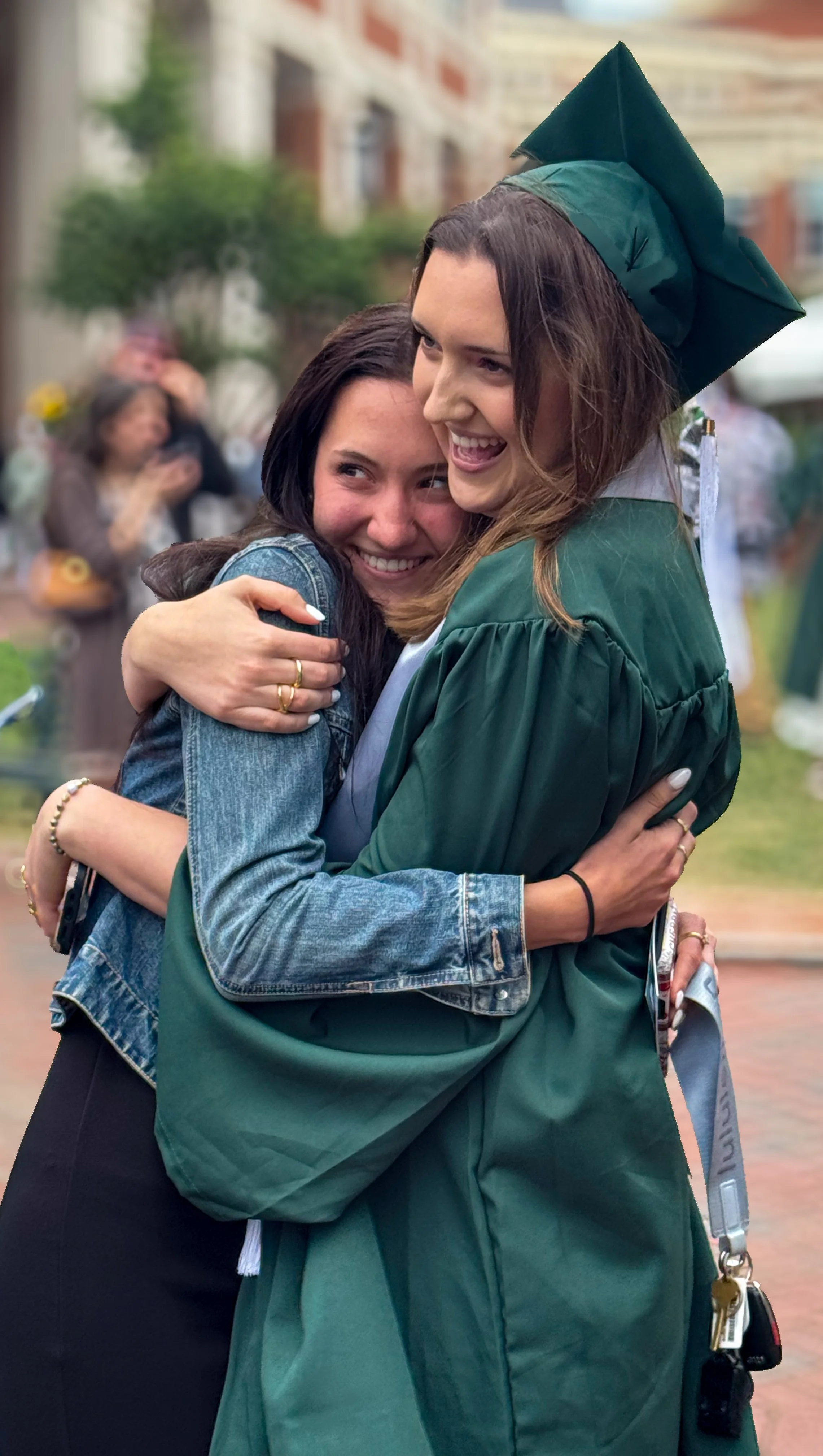 A young woman in a green graduation cap and gown hugging a smiling woman in a denim jacket at an outdoor graduation celebration.