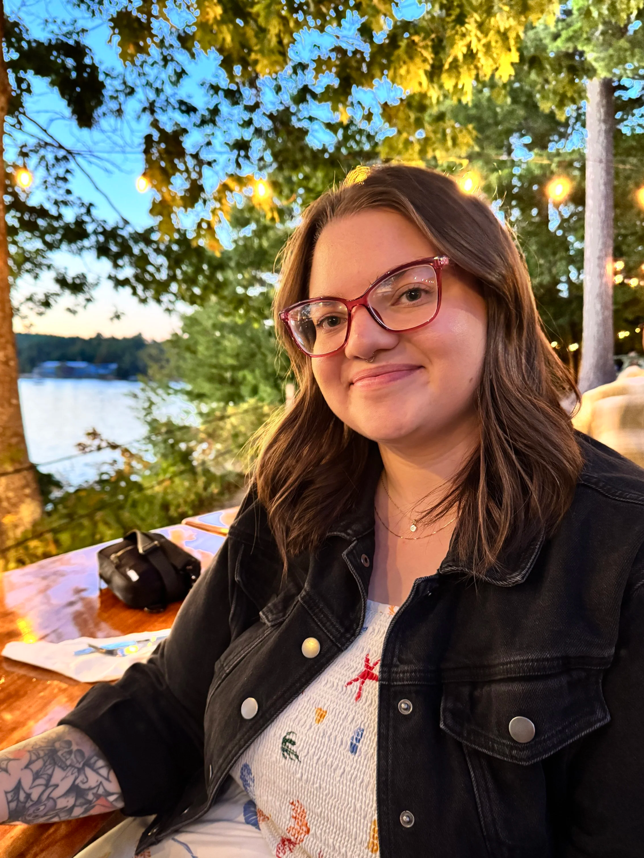 A woman with glasses and shoulder-length wavy hair sits outdoors at a wooden table, with string lights and trees in the background, near a body of water during evening.