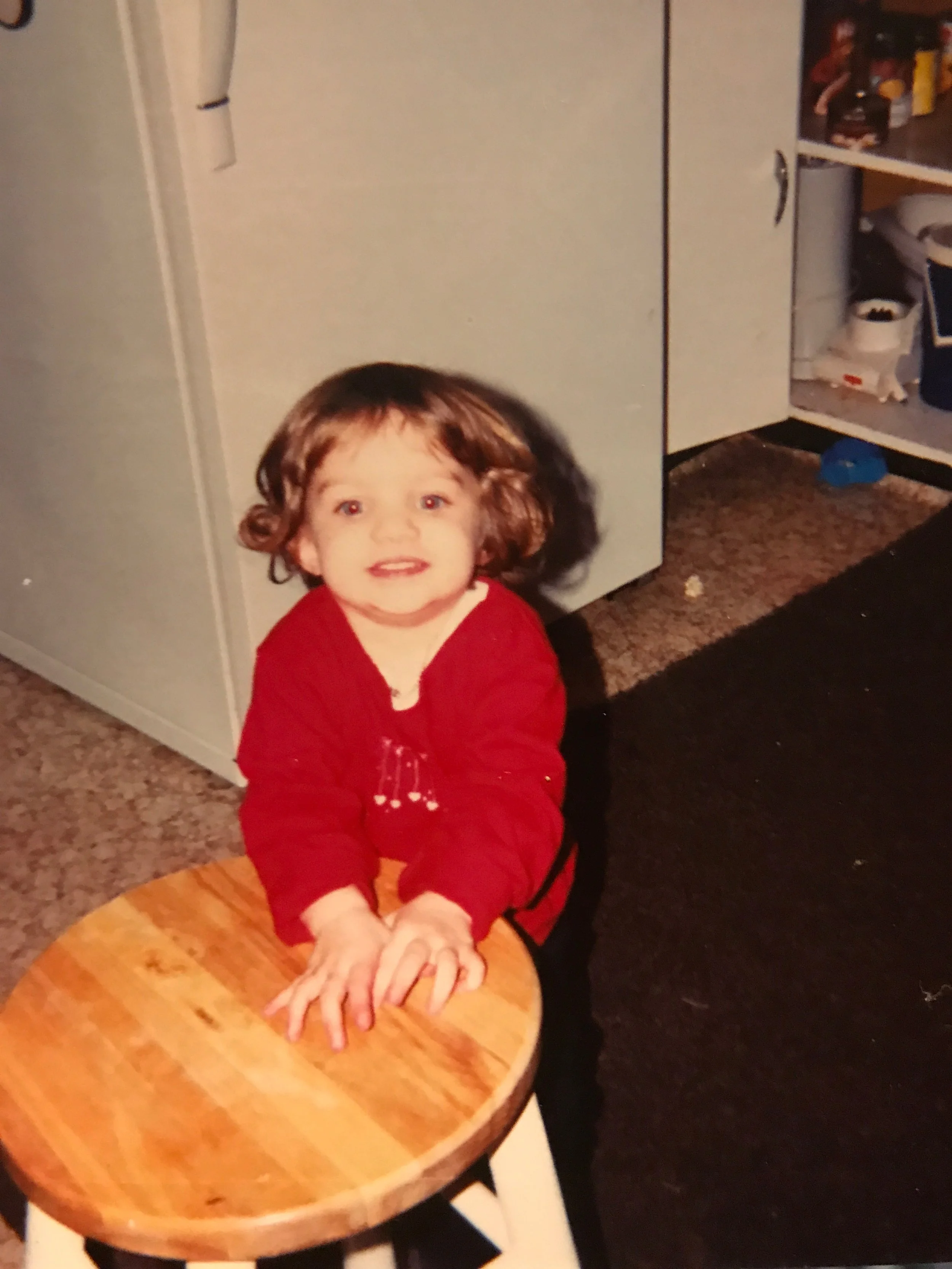 A young girl with brown, curly hair and an excited expression, wearing a red long-sleeve shirt, leaning on a wooden stool with her hands on top, indoors in a room with a beige carpet and cabinets.