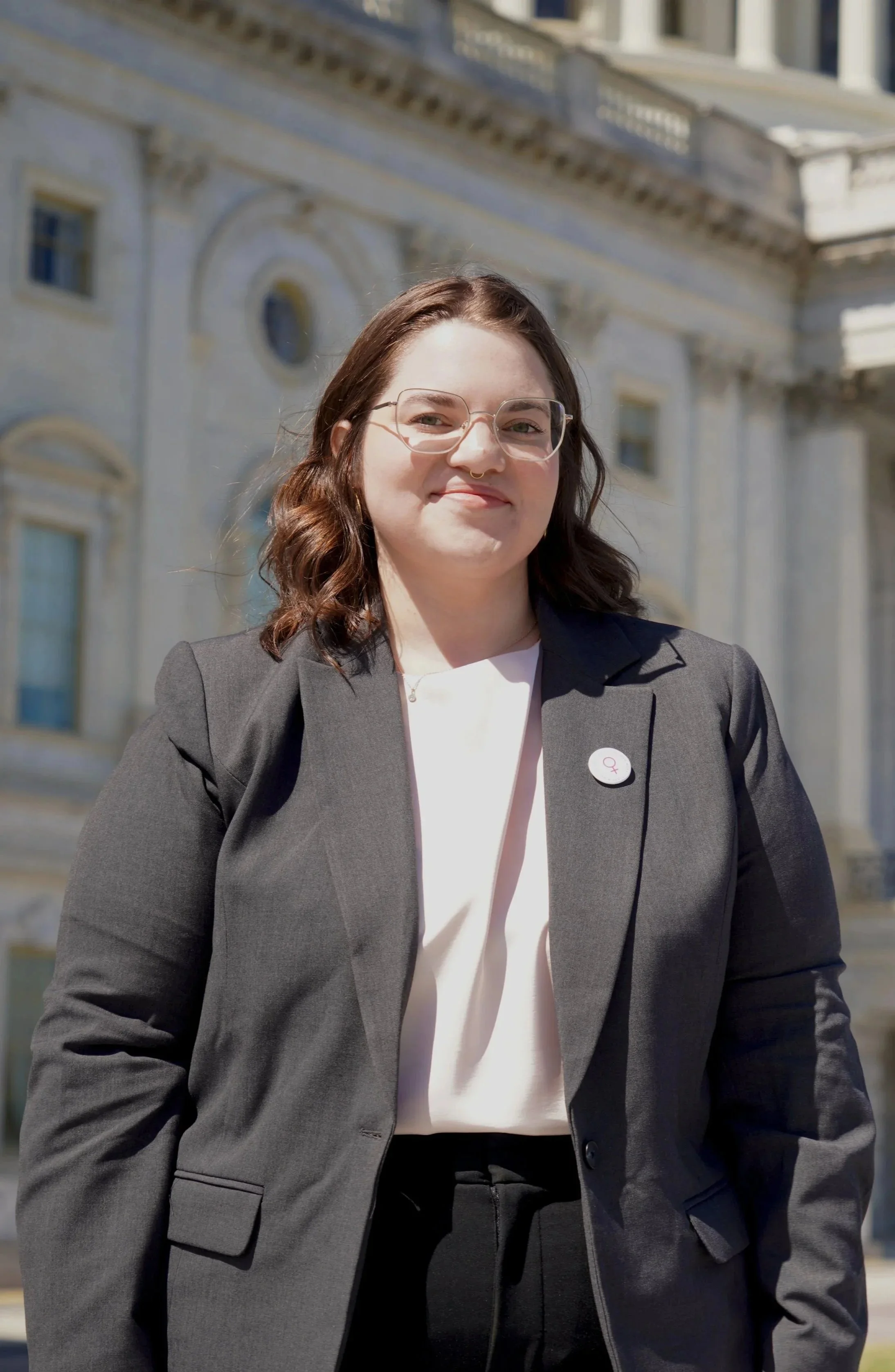 A woman with brown hair, glasses, and a septum piercing, wearing a black blazer and cream blouse, standing outdoors in front of a historic white building.