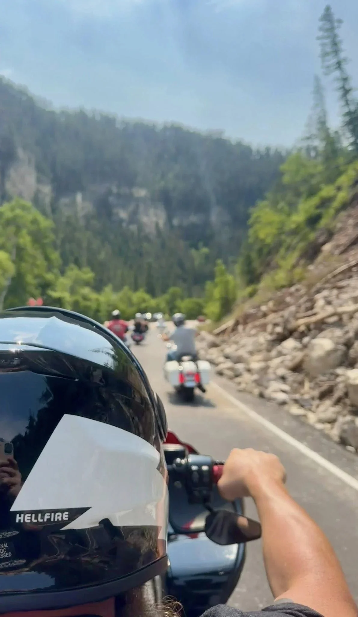 Motorcycle rider wearing a helmet with the brand 'Hellfire' riding on a scenic mountain road with other motorcycles visible in front, surrounded by trees and rocky cliffs.