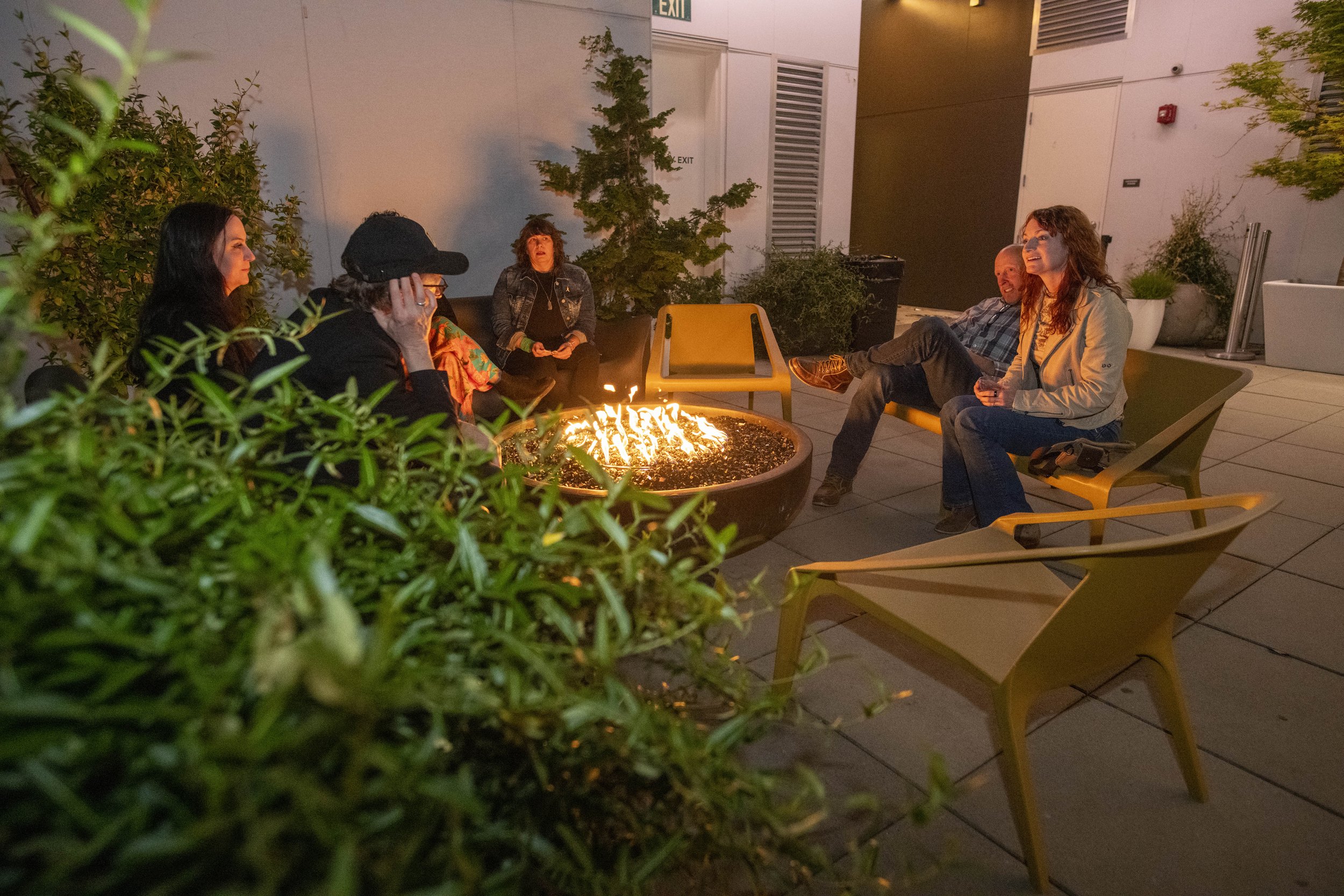Group of five people sitting around a fire pit on a rooftop patio at night, engaging in conversation and enjoying the outdoors.