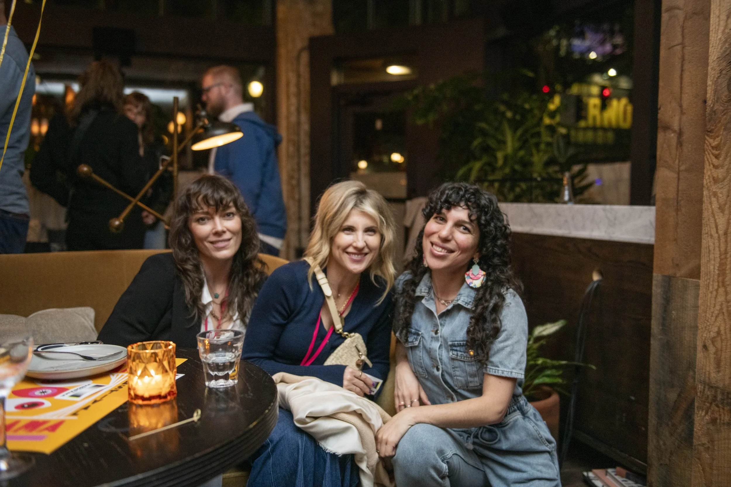 Three people sitting at a table in a restaurant or bar, smiling at the camera, with plates, glasses, and a candle on the table, and other people in the background.
