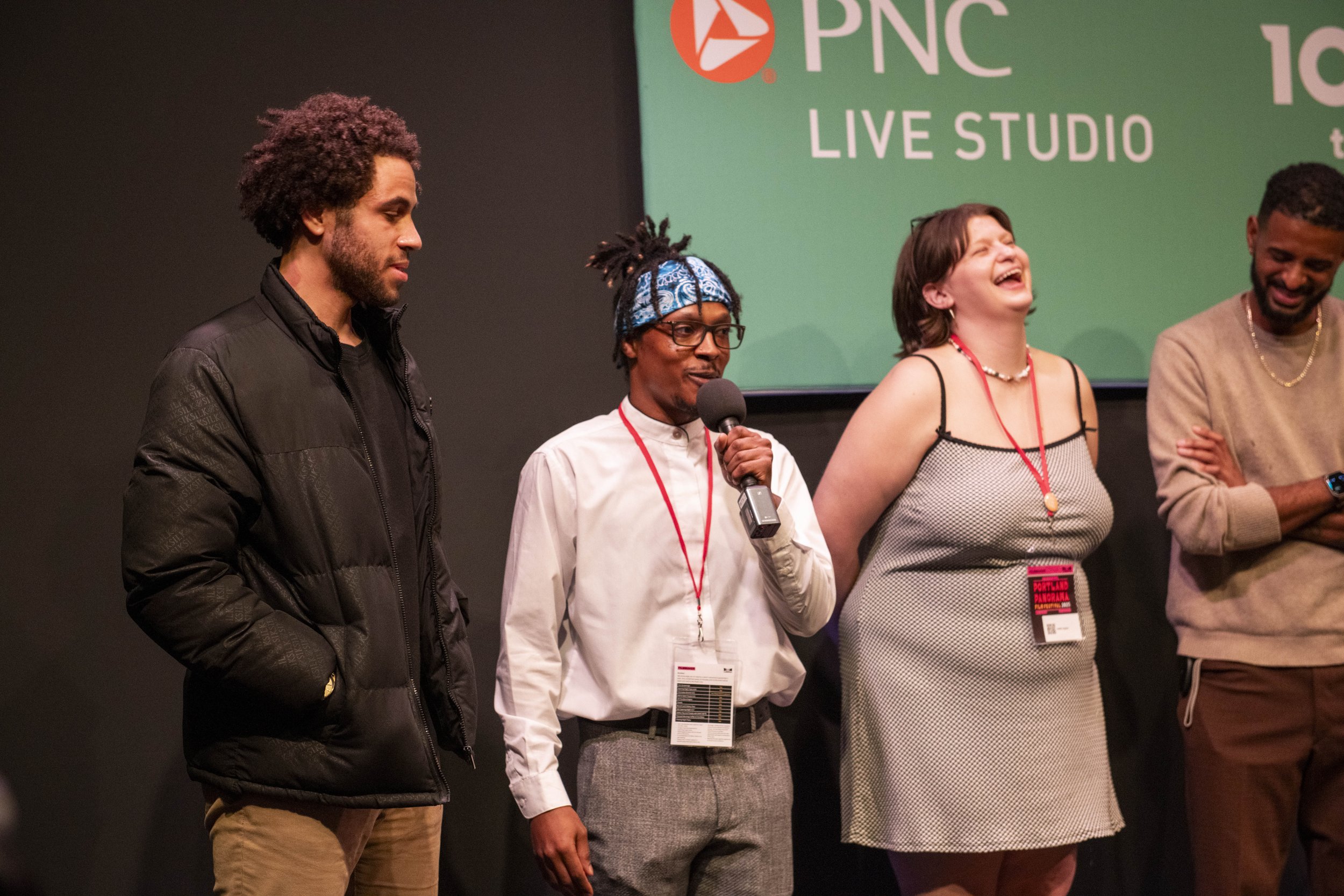A group of five diverse individuals standing on stage at PNC Live Studio, with one person speaking into a microphone, and others appearing happy and engaged.
