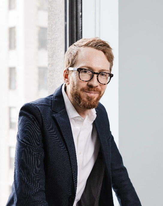 A man with glasses, a beard, and brown hair sitting by a window in an office building.