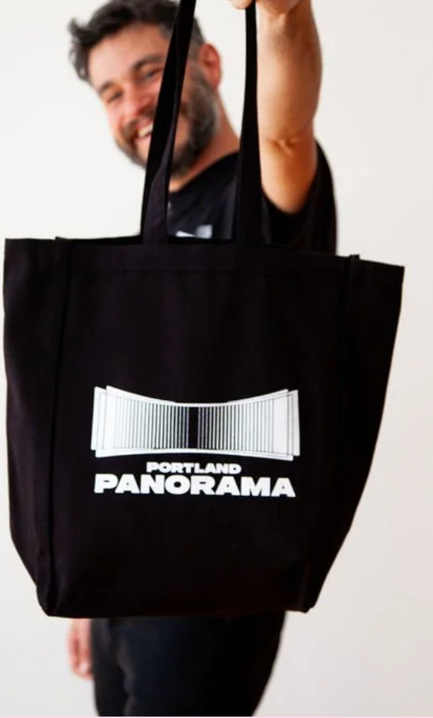 A man with glasses and a beard is smiling and holding up a large black tote bag with the words 'Portland Panorama' and a barcode design printed on it.
