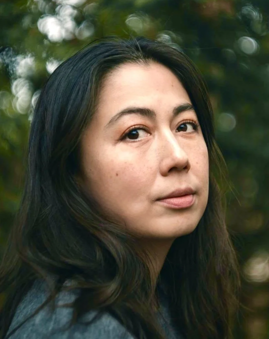 Close-up of a person with long dark hair and tan complexion, standing outdoors with blurred greenery in the background.