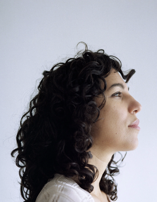 Profile of a woman with dark curly hair looking to the right against a plain light background.