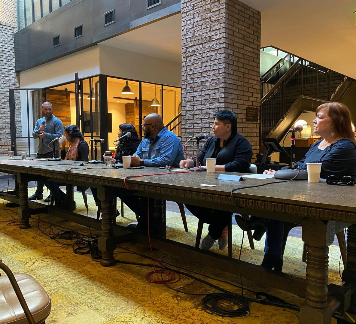 Panel discussion with six people seated at a long table, one person standing with a microphone, in a modern indoor space with brick walls and large glass windows.