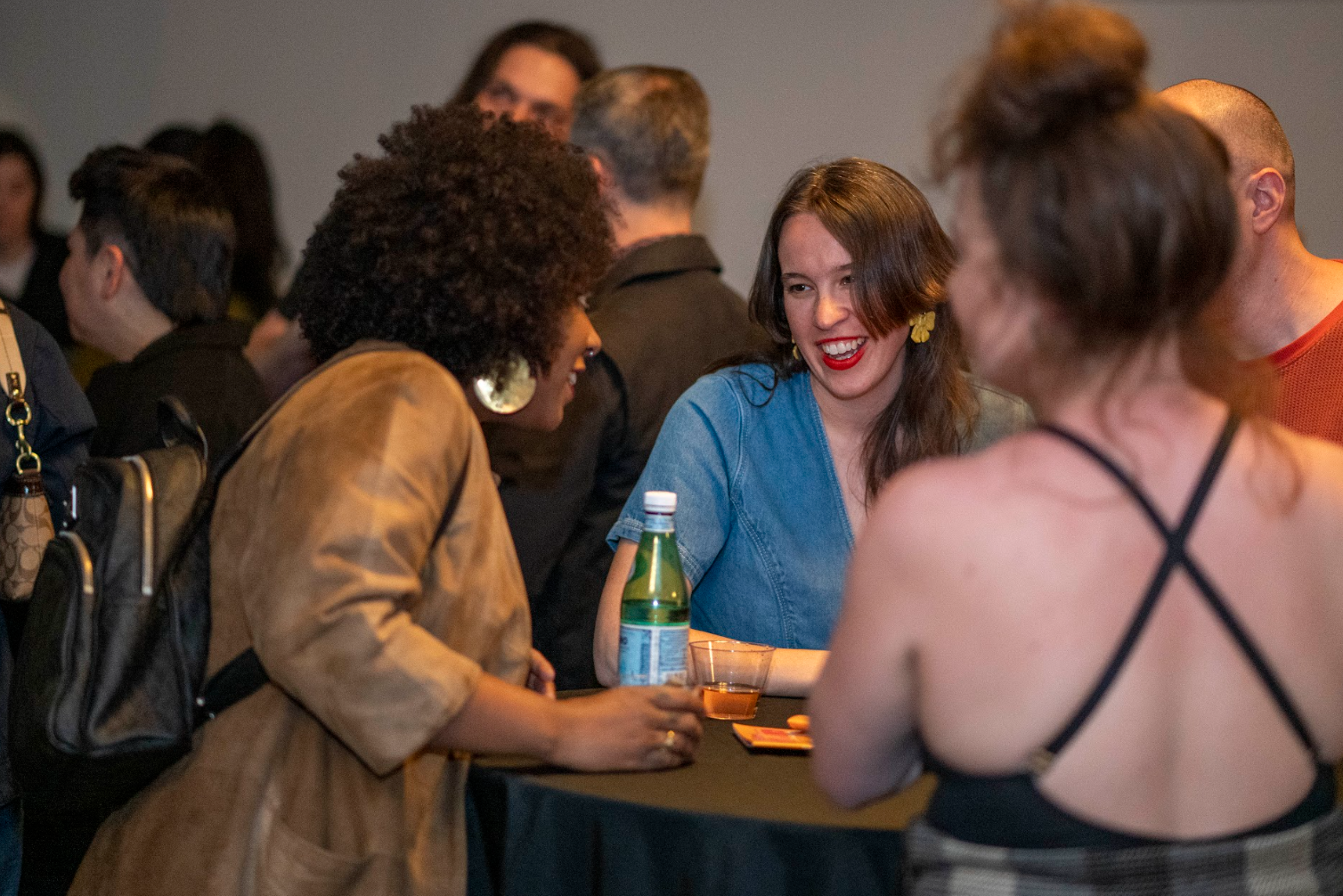 Group of people socializing at a gathering, standing around a table with drinks, engaging in conversation, smiling and enjoying each other's company.