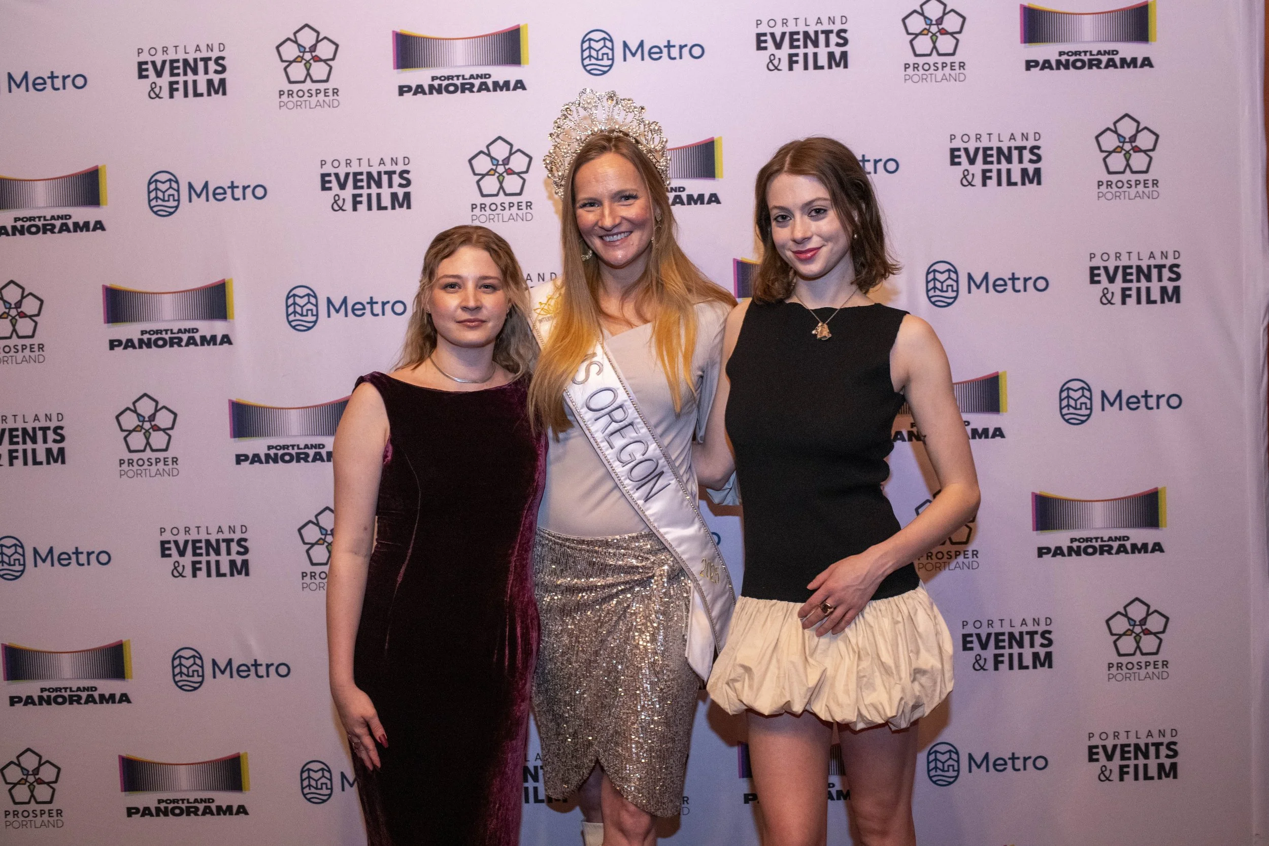 Three people standing in front of a backdrop with Portland Events & Film, Metro, Prosper Portland, and Portland Panorama logos. The person in the middle is wearing a sash and a crown.