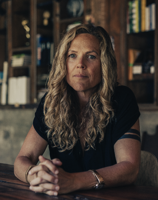 A woman with long, curly blonde hair sitting at a table in a room with wooden bookshelves filled with books and decor.