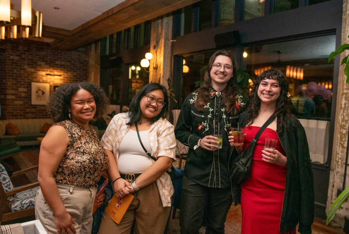 Group of four friends smiling and posing for a photo at a social event in a warmly lit indoor setting, some holding drinks.