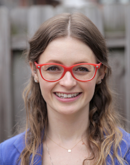 A woman with long wavy brown hair wearing red glasses, a blue top, and small earrings, smiling outdoors in front of a wooden fence.