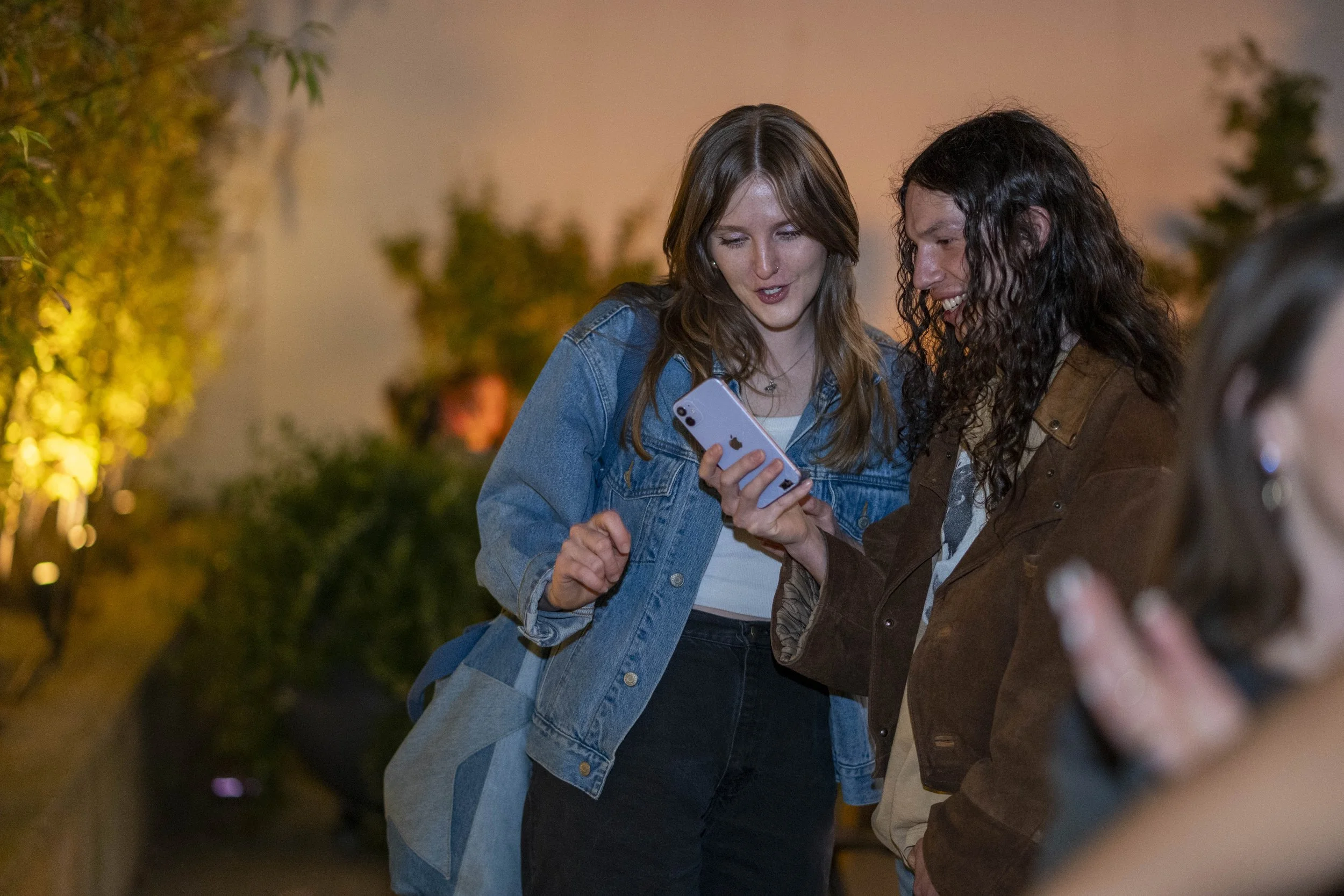 Two young people look at a smartphone together at night outdoors, with warm lighting and blurred background.