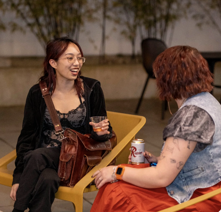 Two people sitting outdoors, chatting and laughing, with drinks in hand, during evening or night time.