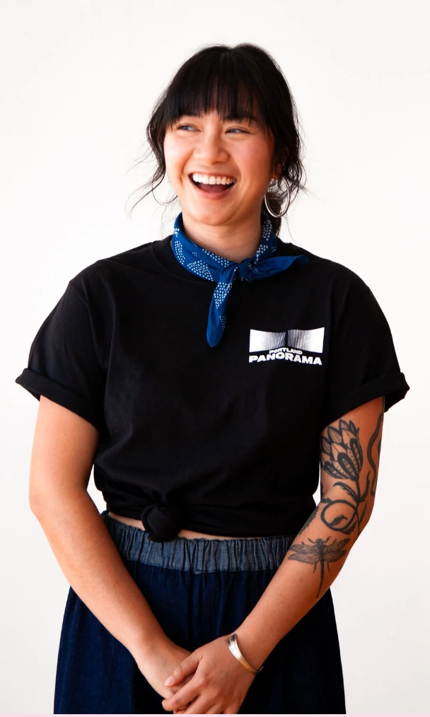 A young woman with black hair and bangs, wearing a black t-shirt with 'Portland Panorama' logo, a blue bandana around her neck, and dark pants, smiling and looking to her left.