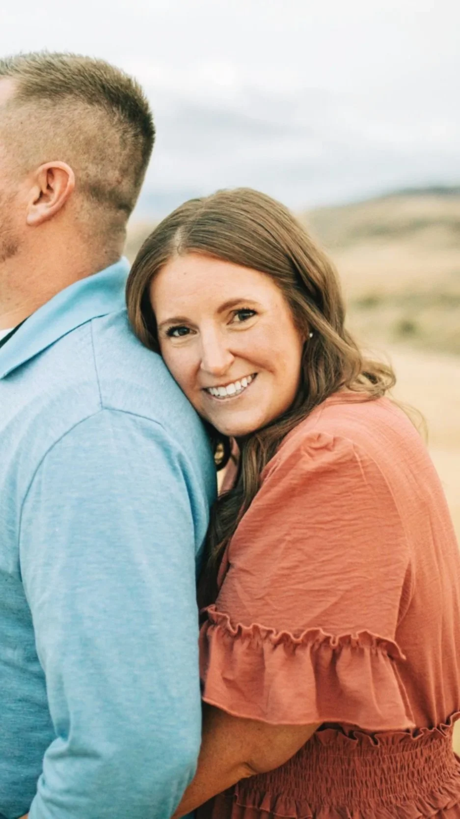 A woman with brown hair smiling, embracing a man wearing a light blue shirt. They are outdoors with a blurred landscape background.