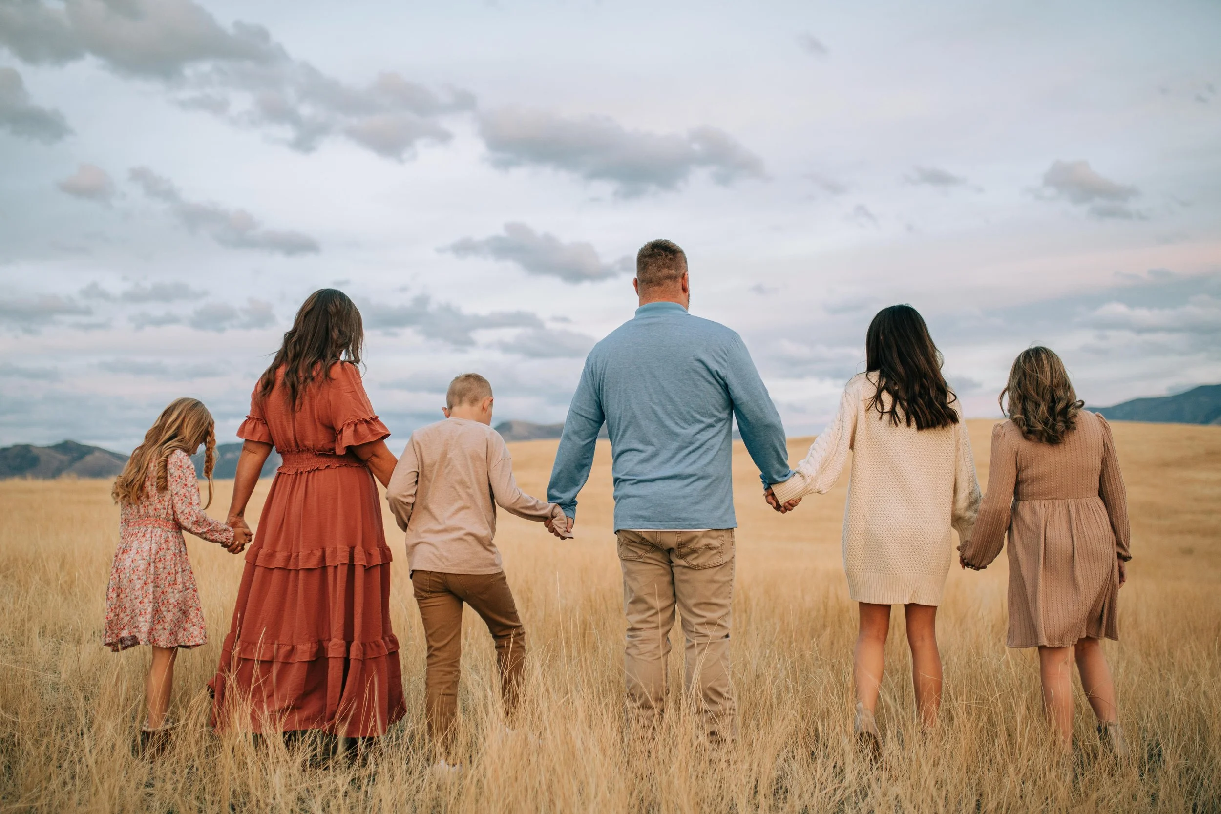 A family holding hands walking in a grassy field with mountains and cloudy sky in the background.