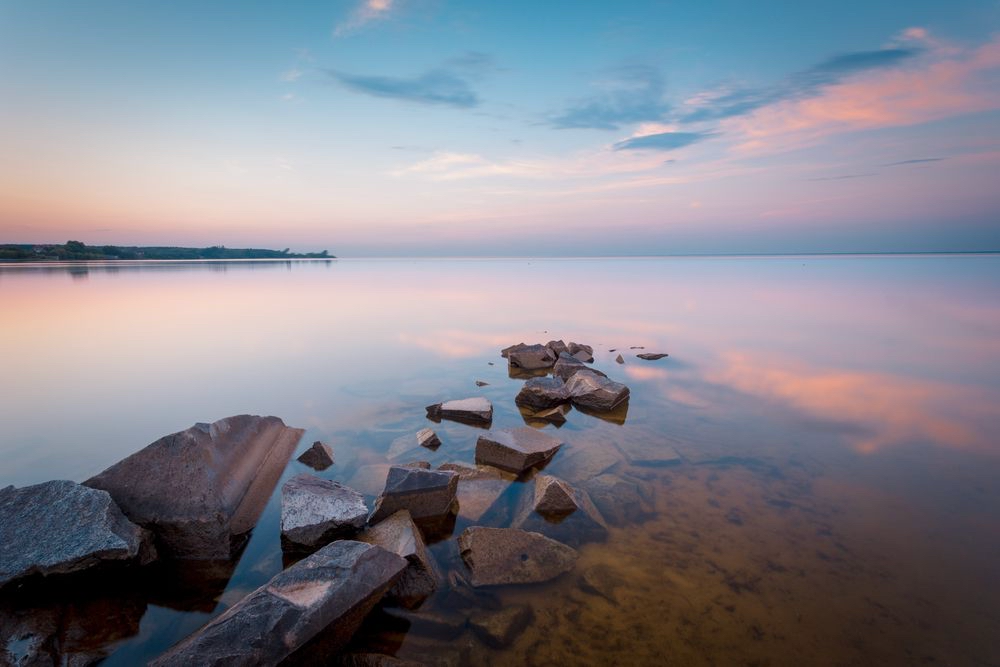 Calm lake at sunset with rocks in the foreground and pink and blue sky reflected in the water