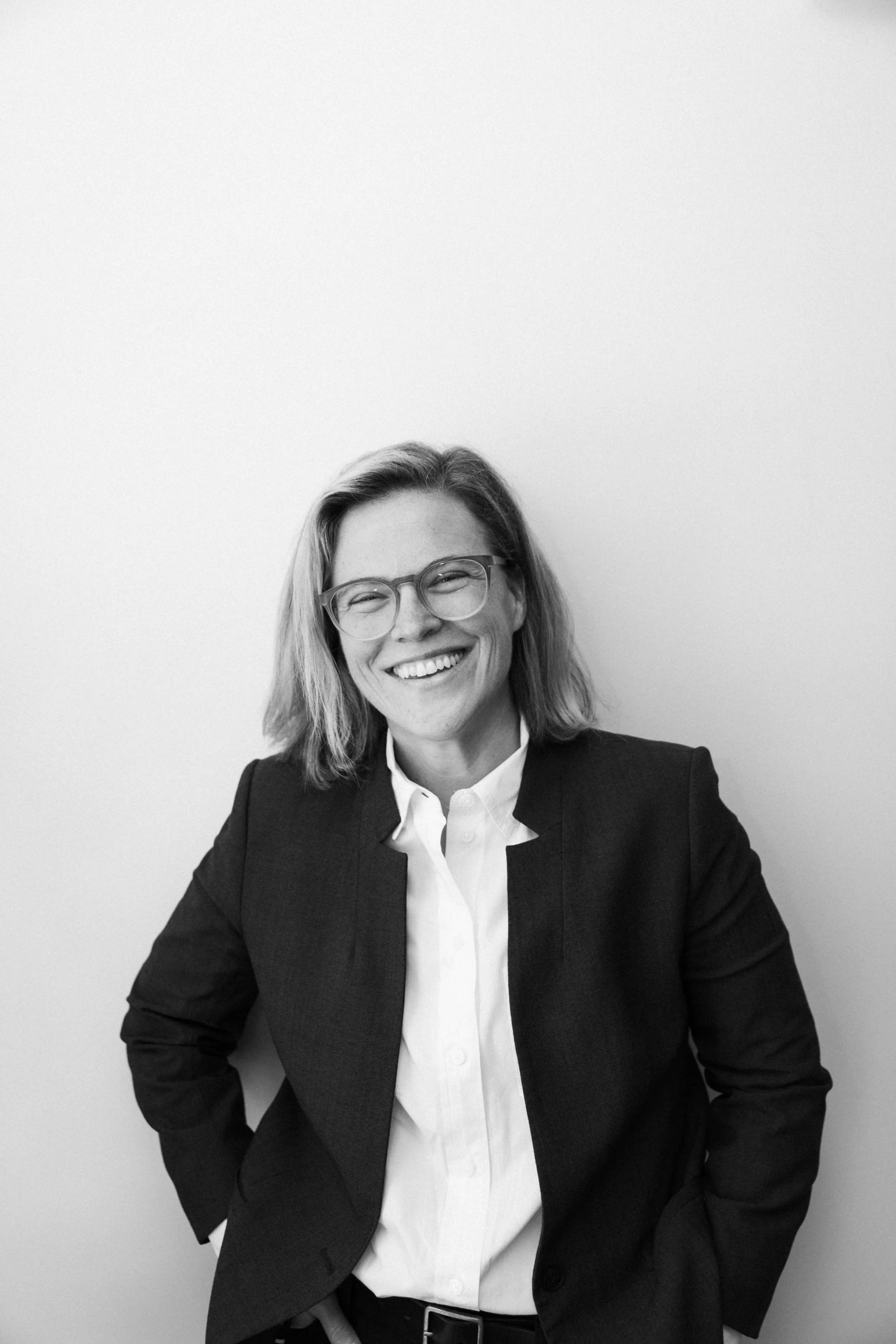 Black and white photo of a woman smiling, wearing glasses, a white button-up shirt, and a dark blazer standing against a plain wall.