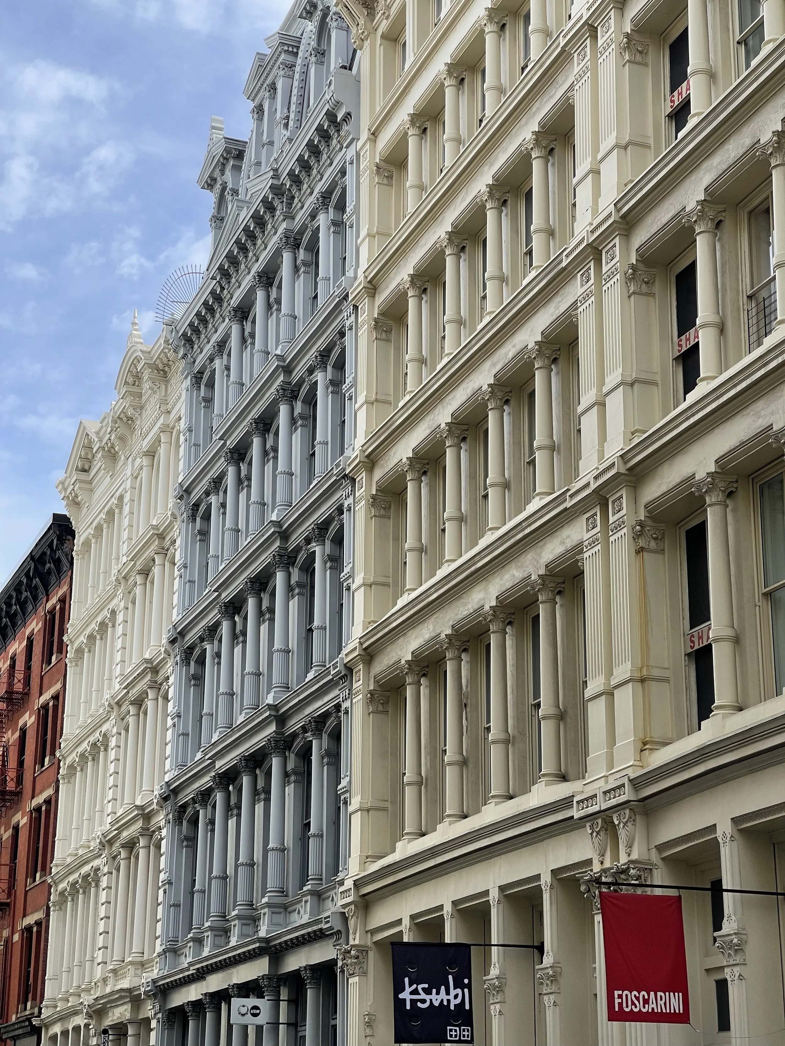 Multiple historic buildings with ornate facades in a city street, featuring decorative columns, large windows, and banners for shops.