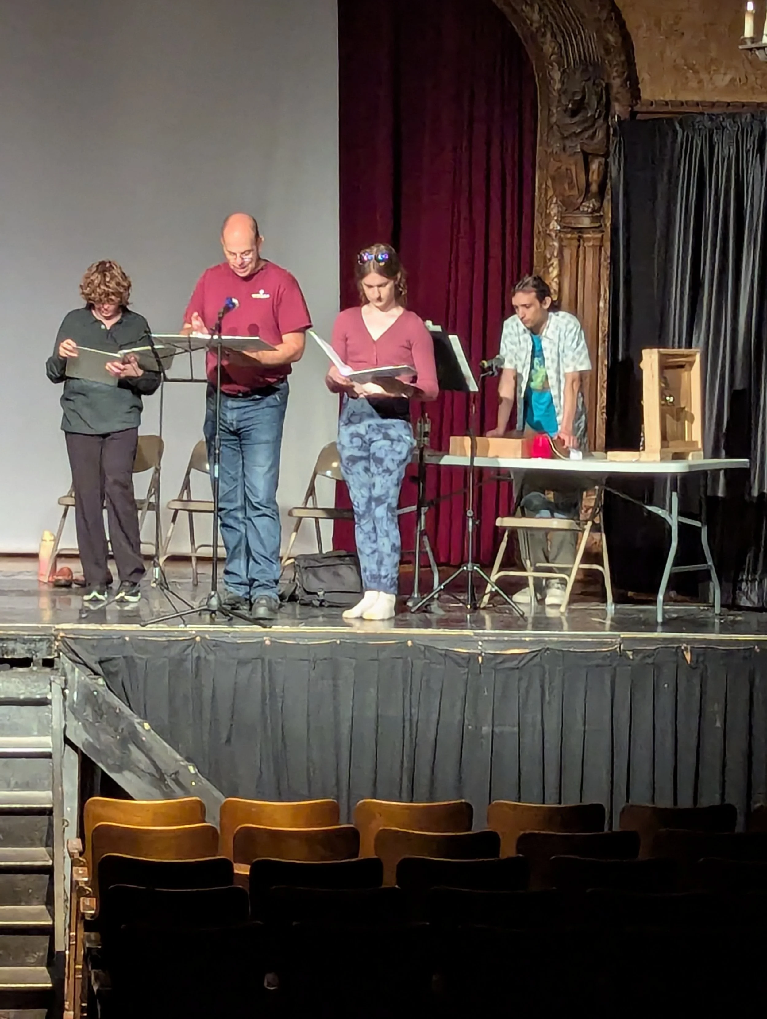 Four people on a stage appear to be rehearsing or practicing, with scripts in hand. They are standing in front of a red curtain, and there are chairs and equipment around them.