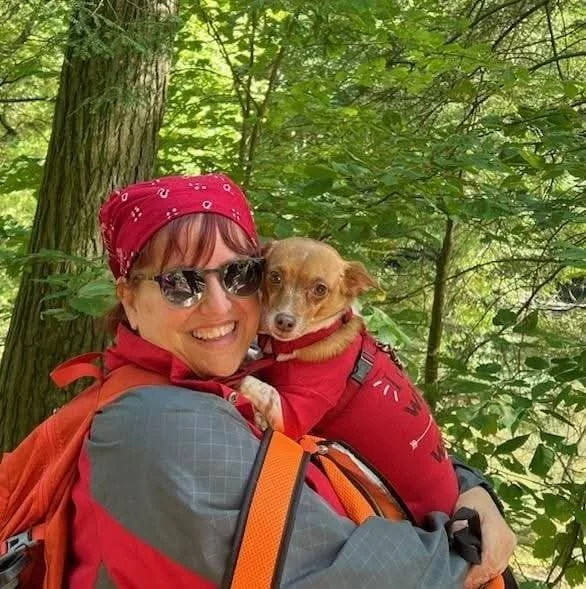 Woman wearing sunglasses and a red bandana, smiling while holding a small dog. The dog is wearing a red outfit and is cuddled close to the woman. They are outdoors in a green wooded area.