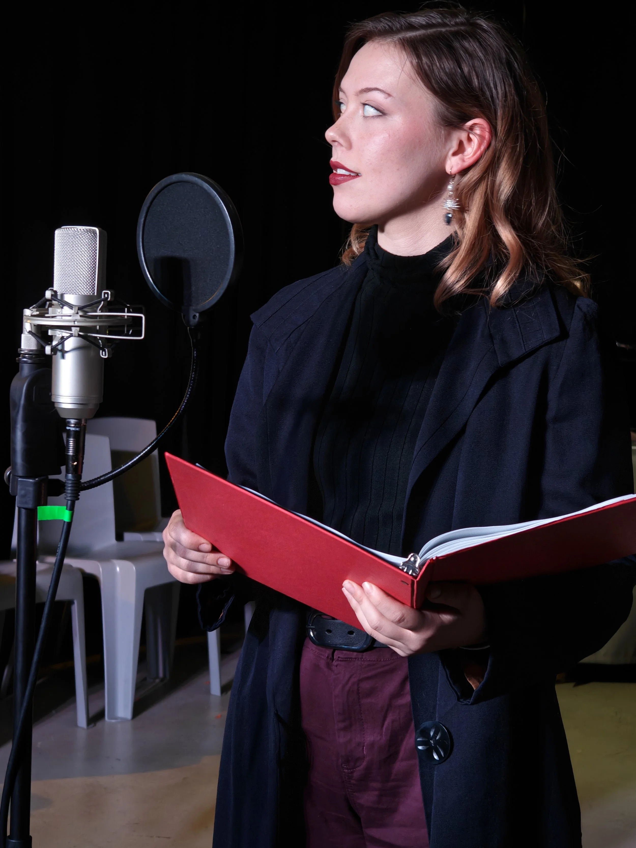 A woman wearing a black jacket, holding a red script folder, standing in a recording studio near a microphone with a pop filter.