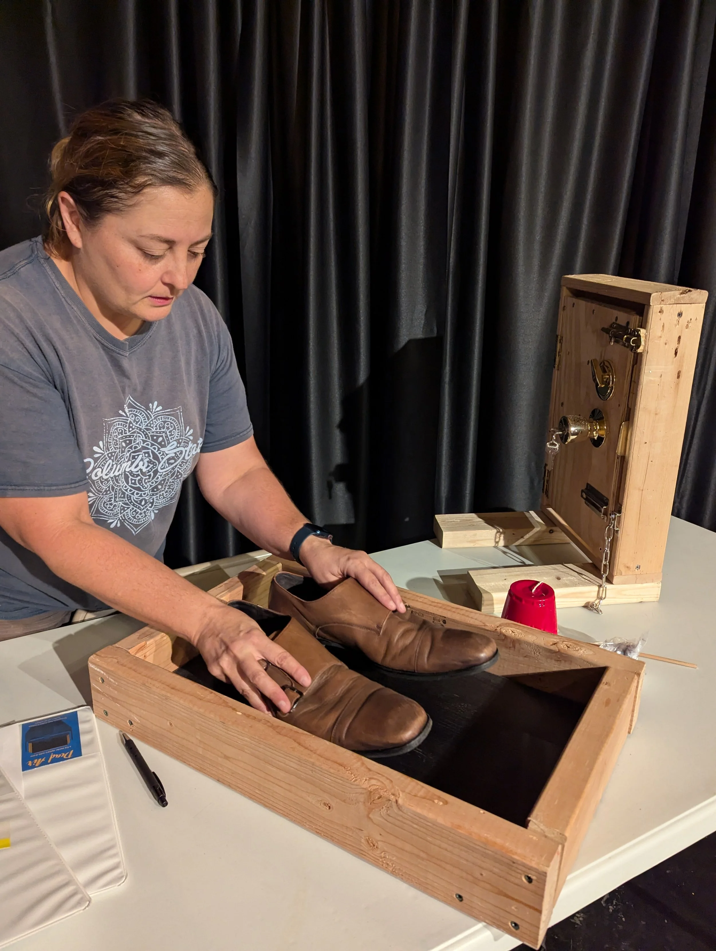 A woman standing at a table with a wooden box containing a pair of brown leather shoes, with a shoe-pressing machine with chains and a latch in the background.