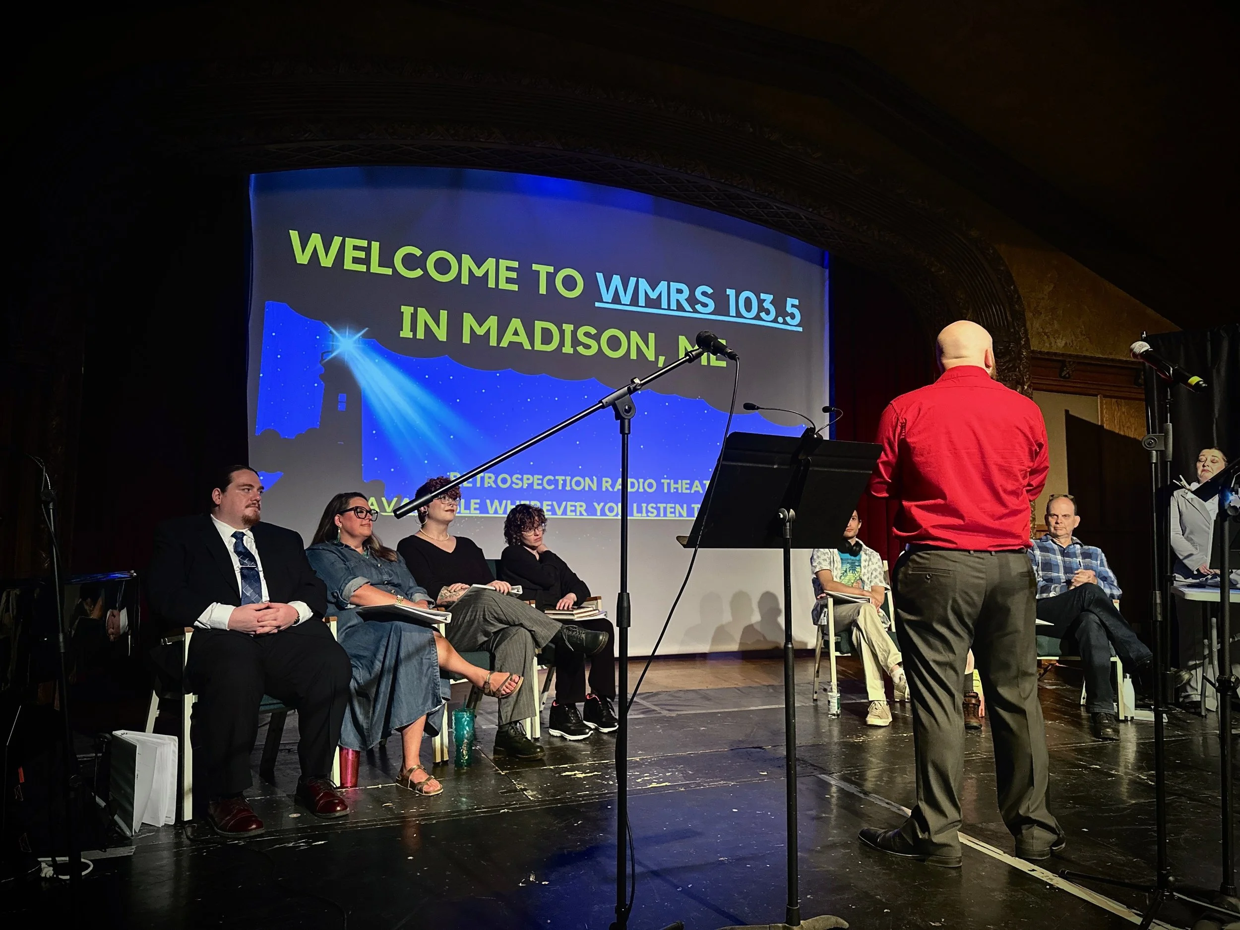 A panel of six people seated on stage facing a man in a red shirt who is speaking. A large screen behind them displays a welcome message to WMRS 103.5 in Madison, Maine, with a graphic of a lighthouse and night sky.
