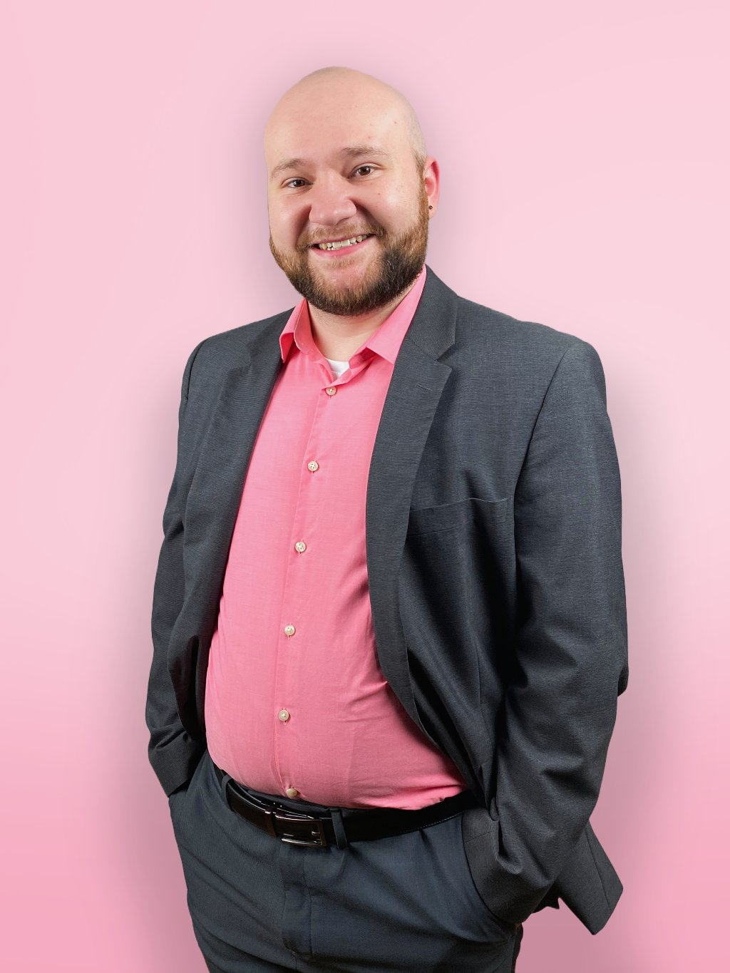 A smiling man with a beard, in a gray suit and pink shirt, standing against a pink background.