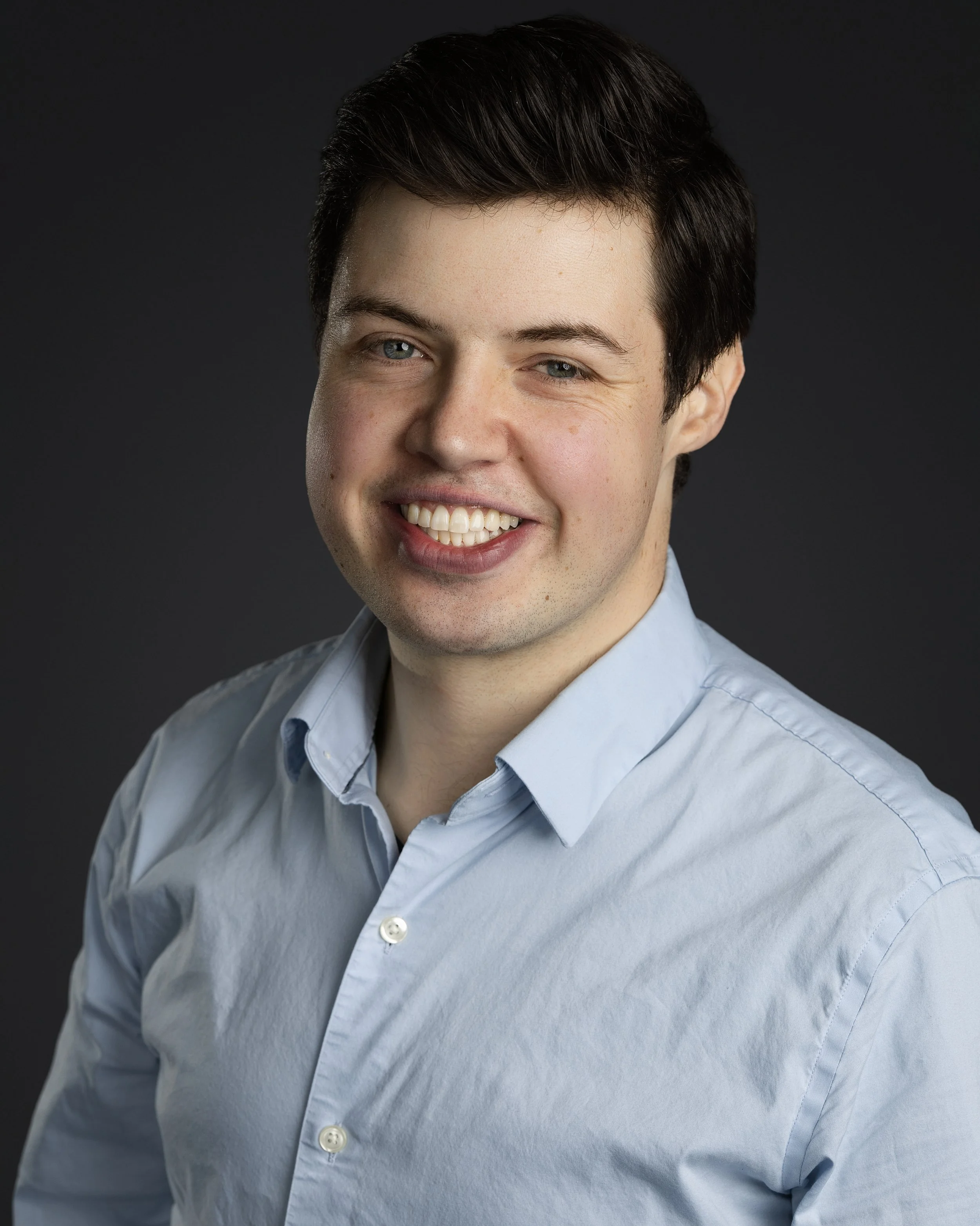 A headshot of a young man with dark hair, smiling, wearing a light blue collared shirt, against a dark background.