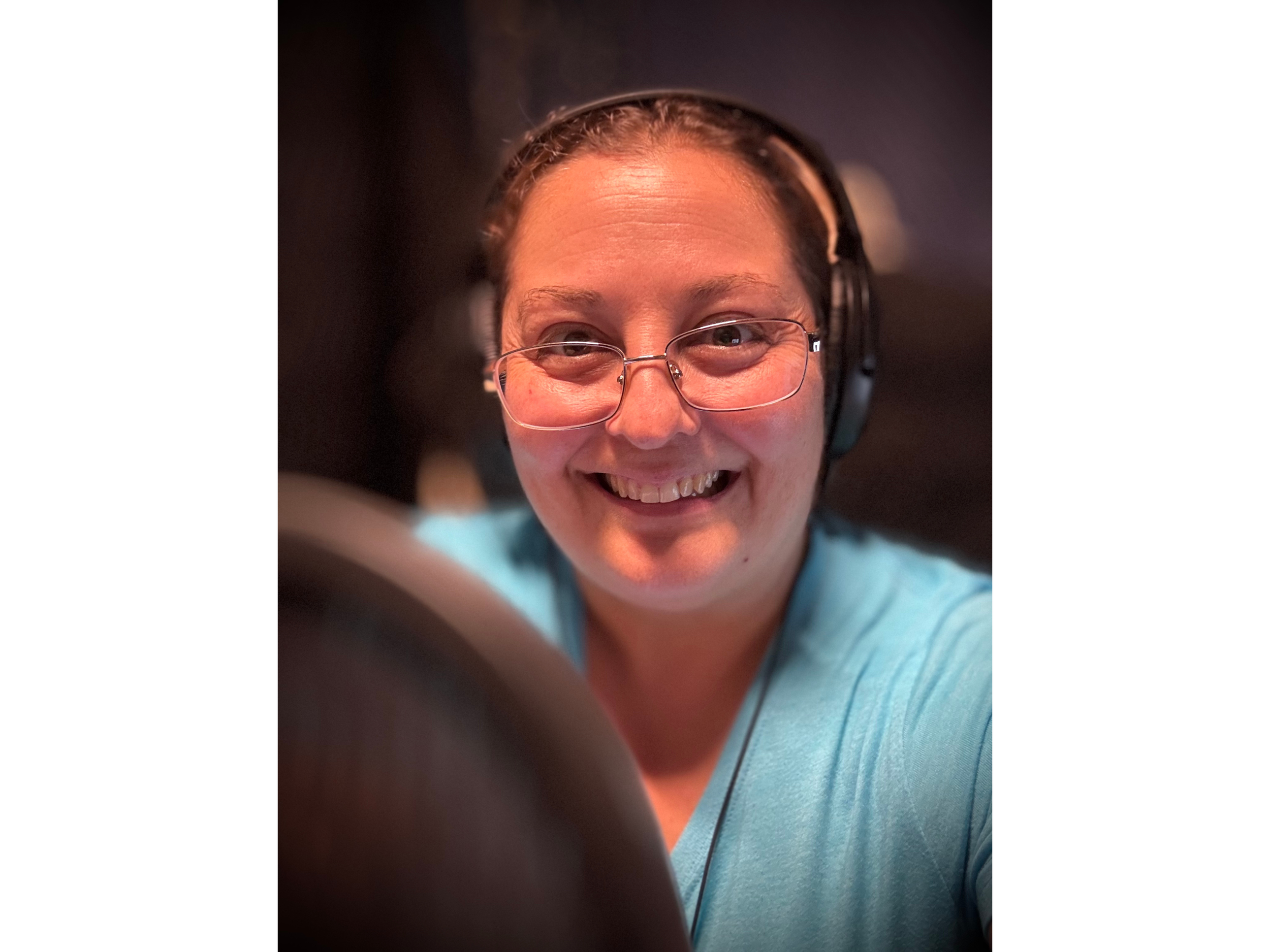 A smiling woman with glasses and headphones taking a selfie, with a dark background and a blurred object in the foreground.