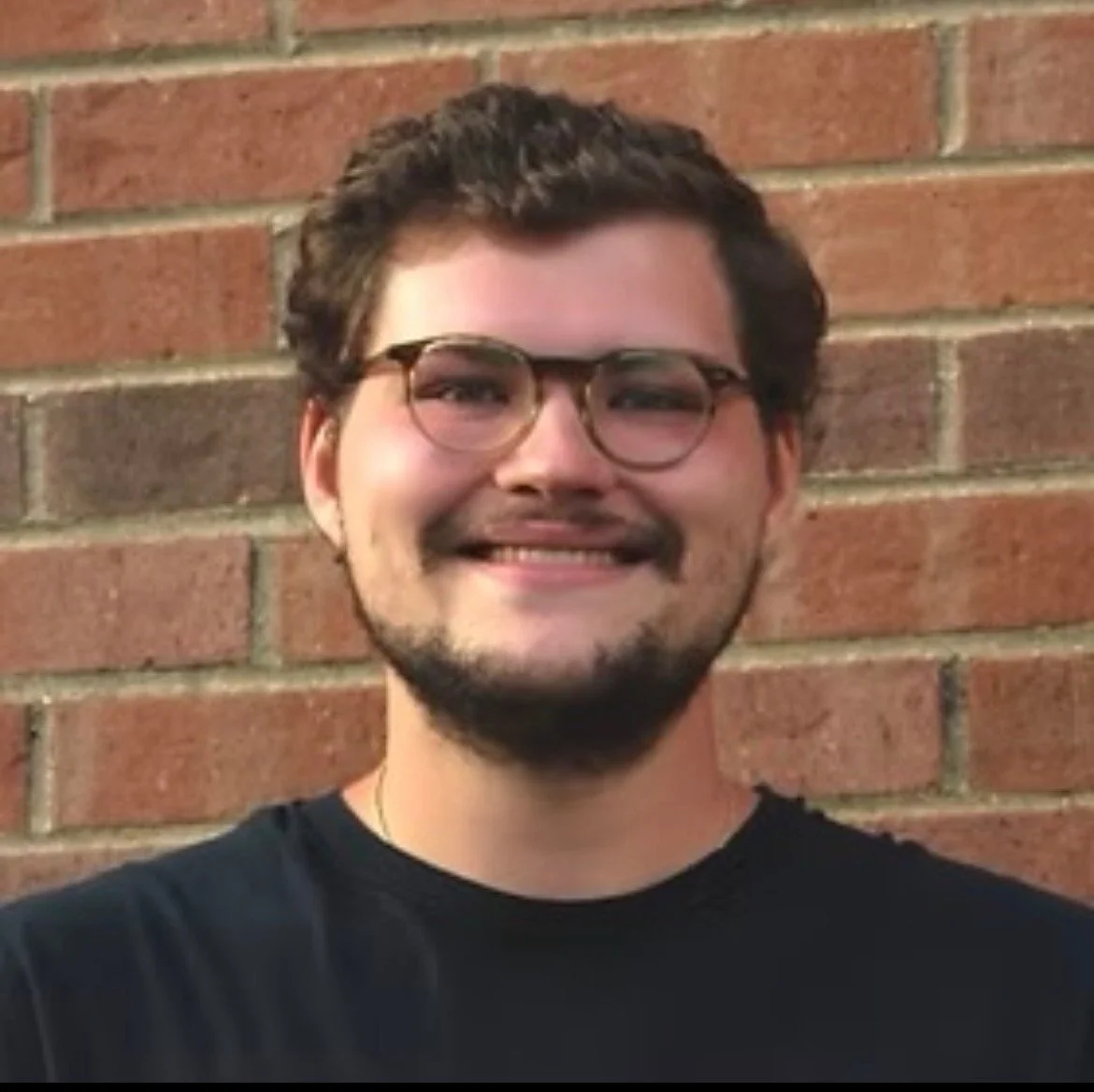 A smiling young man with glasses, a beard, and short curly hair standing against a brick wall.