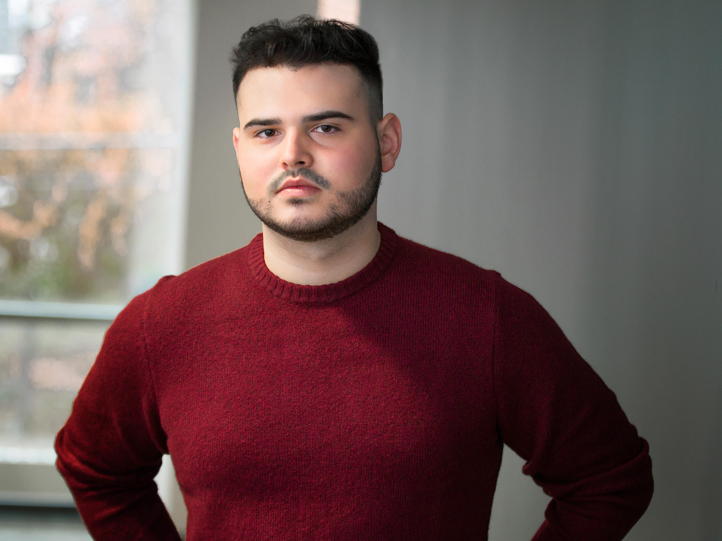 A young man with dark hair, a short beard, and wearing a red sweater, standing indoors near a window with a blurred outdoor view.