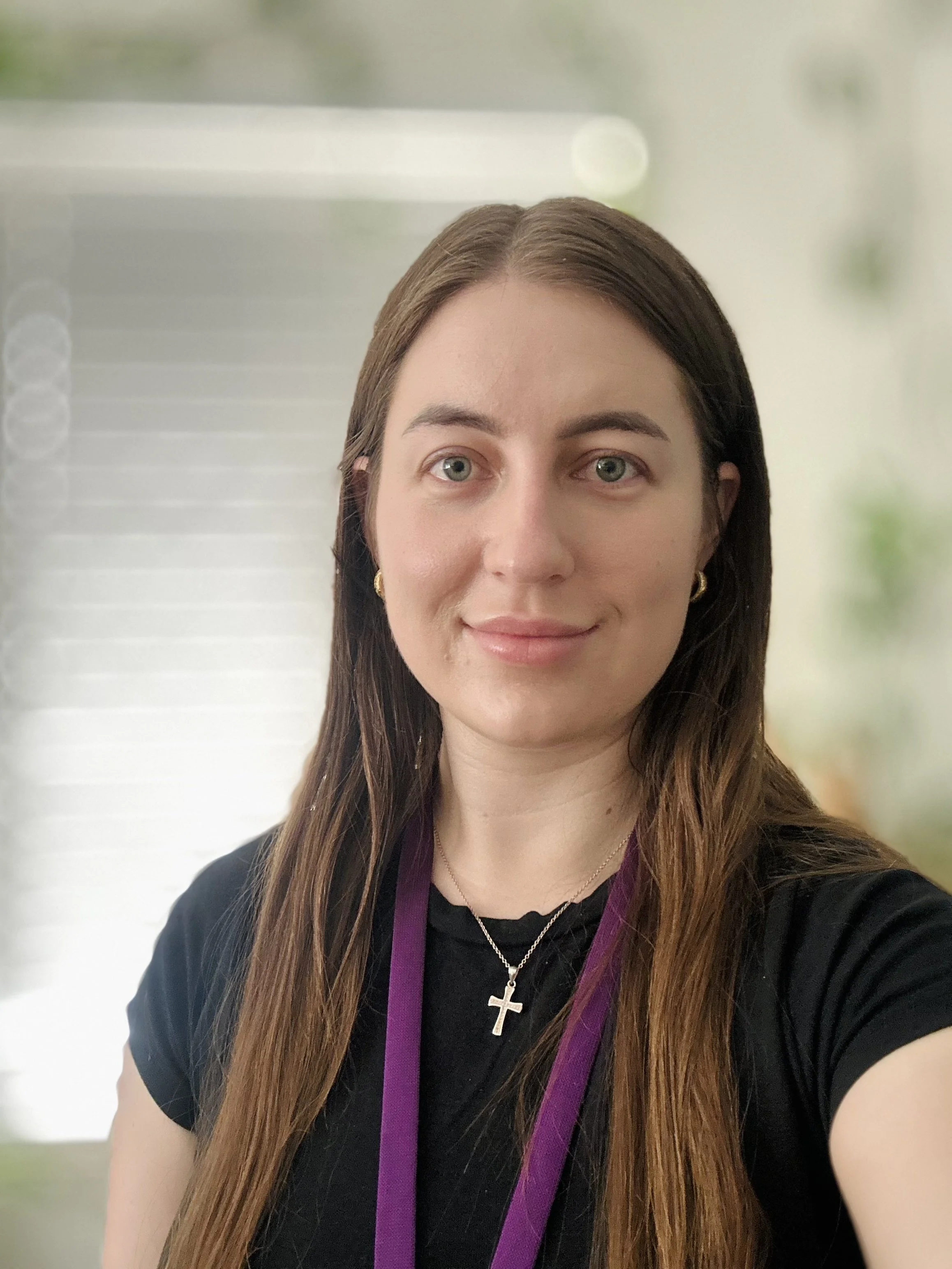 A woman with long brown hair and green eyes smiling, wearing a black shirt, gold earrings, a silver cross necklace, and a purple lanyard, standing indoors with a blurred background.