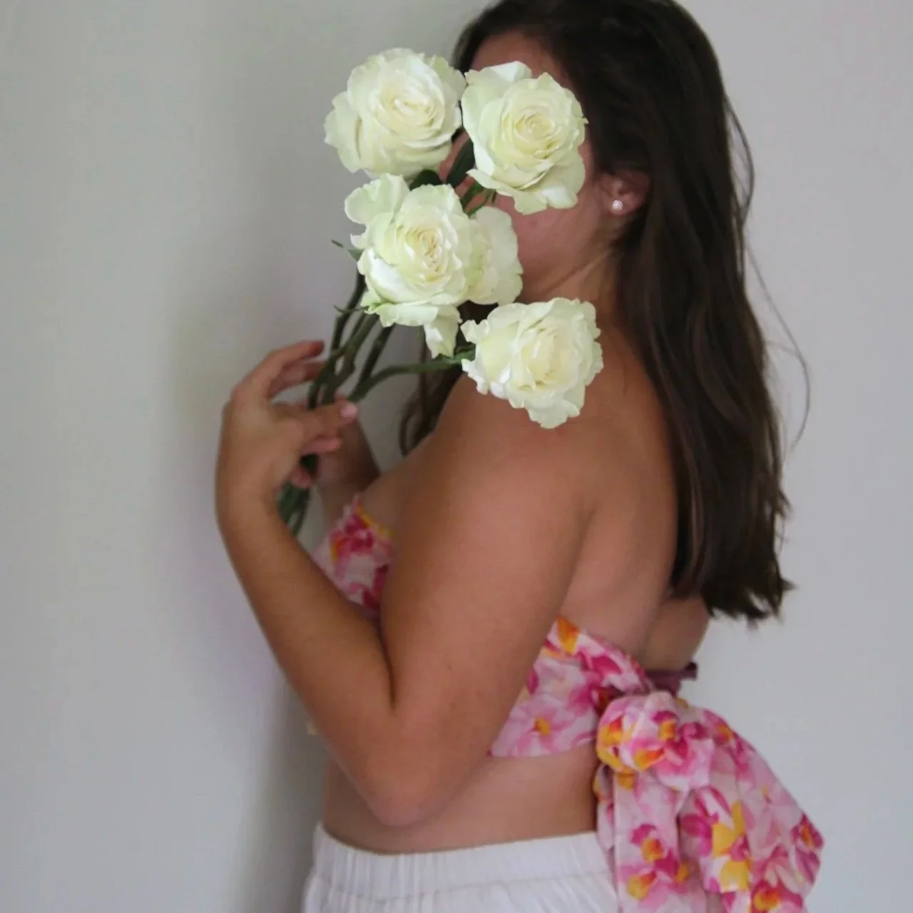 A woman with long dark hair is holding a bouquet of large white roses in front of her face. She is wearing a colorful floral top and white pants, standing against a plain light-colored background.