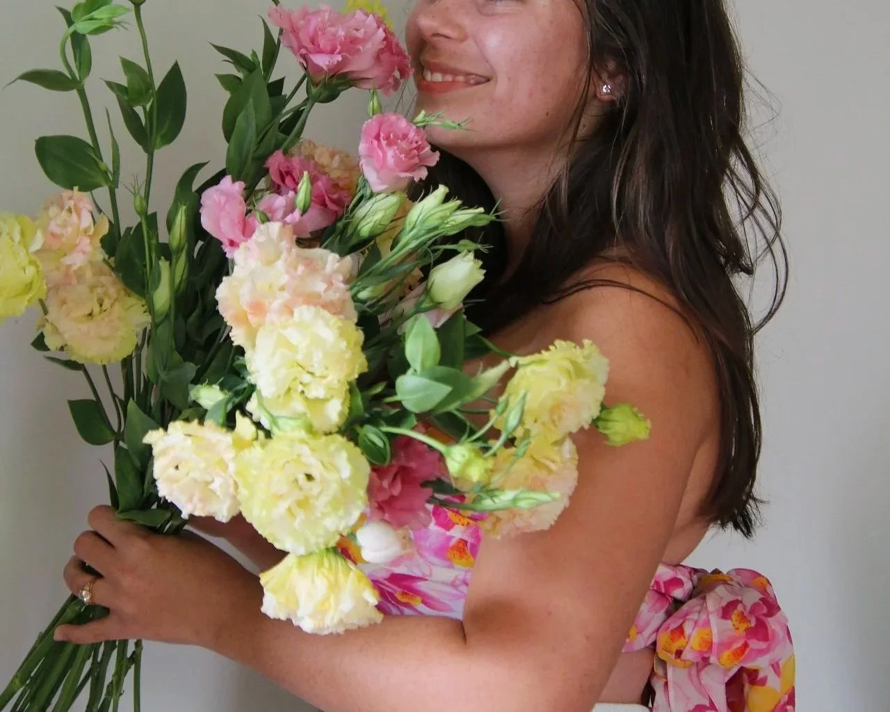 Woman holding a large bouquet of pink, yellow, and white flowers, smiling with dark, wavy hair, wearing a colorful dress with a floral pattern and a pink bow at the back.