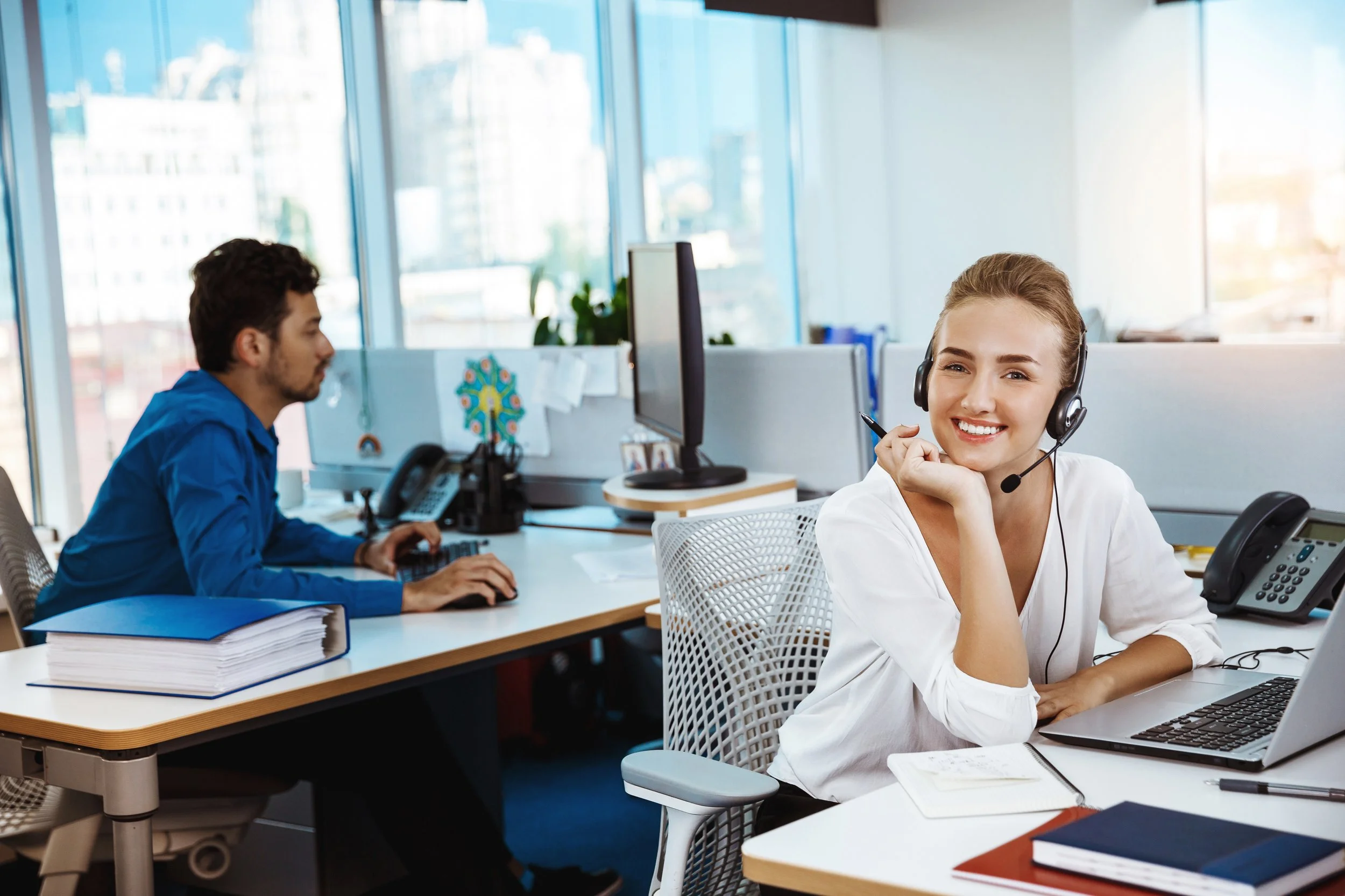 A woman with a headset smiling at her desk in an office setting, with a man working at his computer in the background.