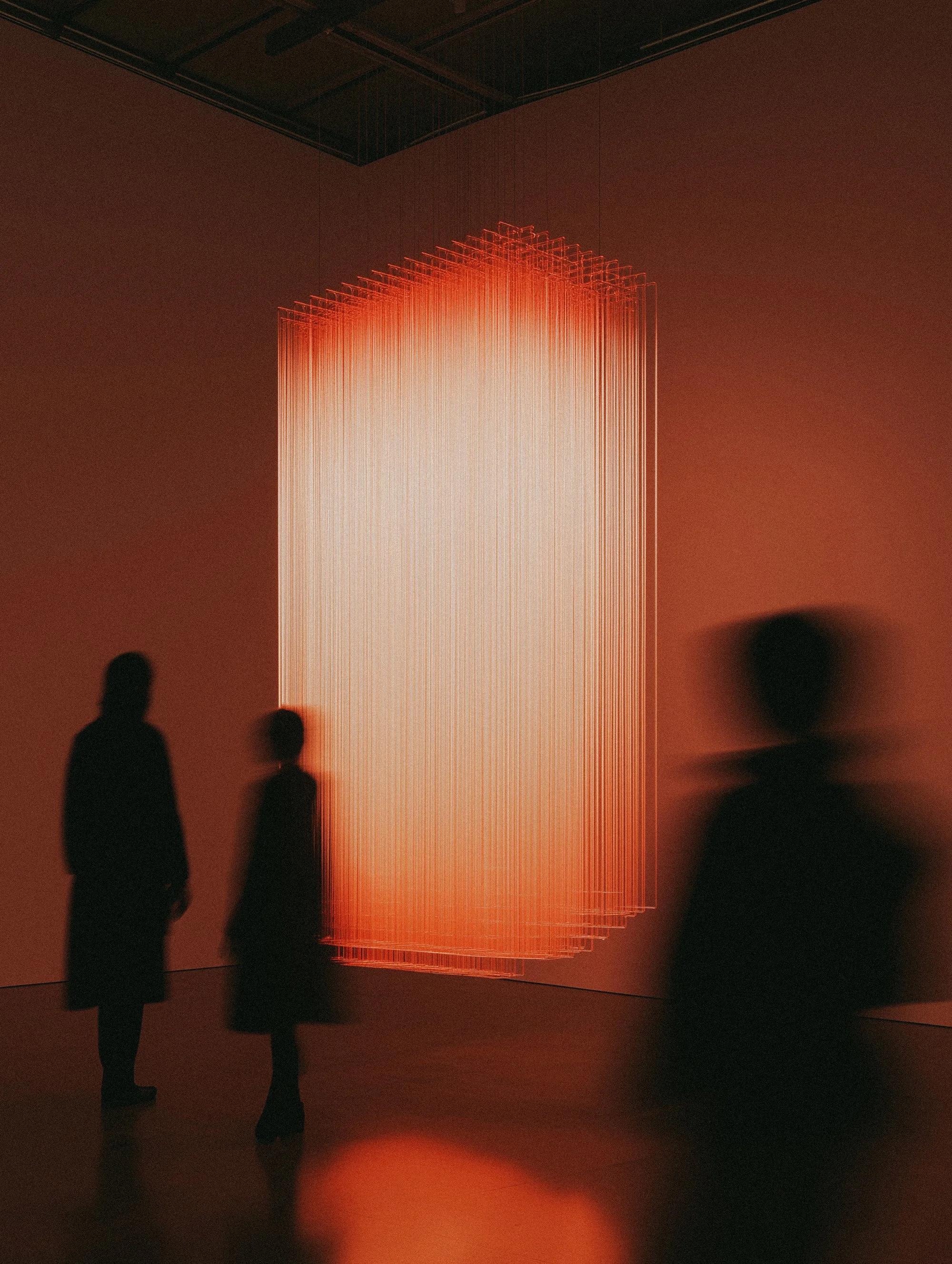 Silhouetted people viewing an art installation made of glowing vertical strings in a dimly lit gallery.