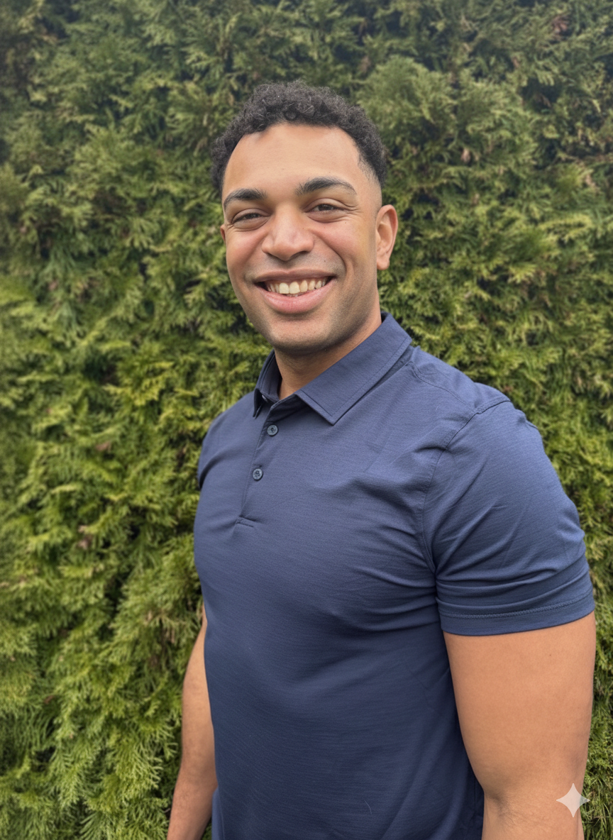 A smiling man with short curly hair wearing a navy blue polo shirt standing outdoors in front of green foliage.