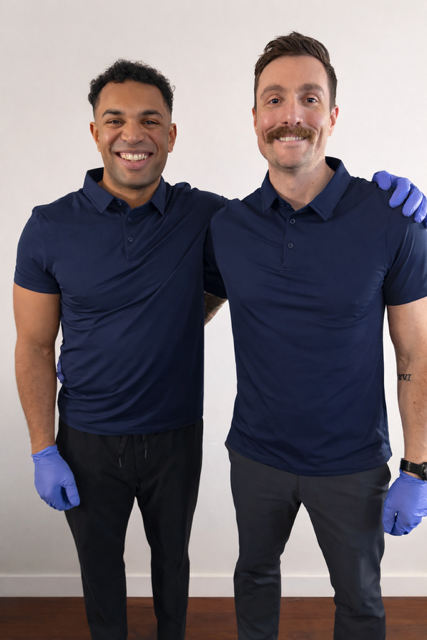 Two men standing side by side, smiling at the camera, wearing navy blue polo shirts, black pants, and blue gloves, in front of a plain white wall.