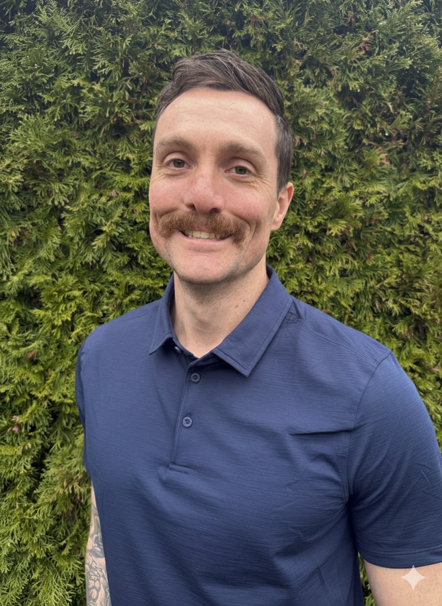 A man with short dark hair, a mustache, and light skin, smiling outdoors in front of a lush green bush, wearing a navy blue polo shirt.