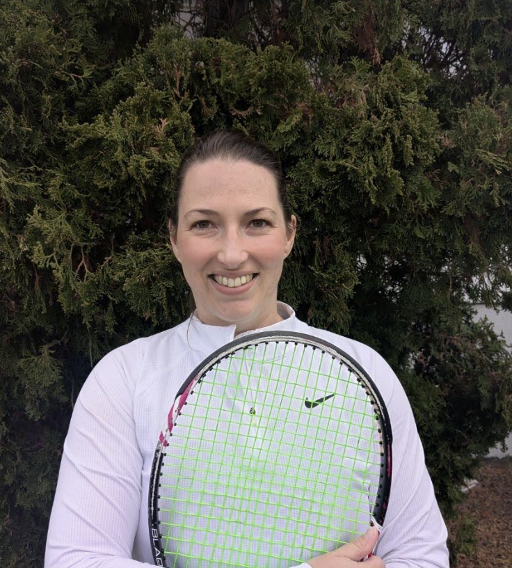 A woman, Kimberley Hartwig, smiling outdoors, holding a tennis racquet, standing in front of a large green bush or tree.