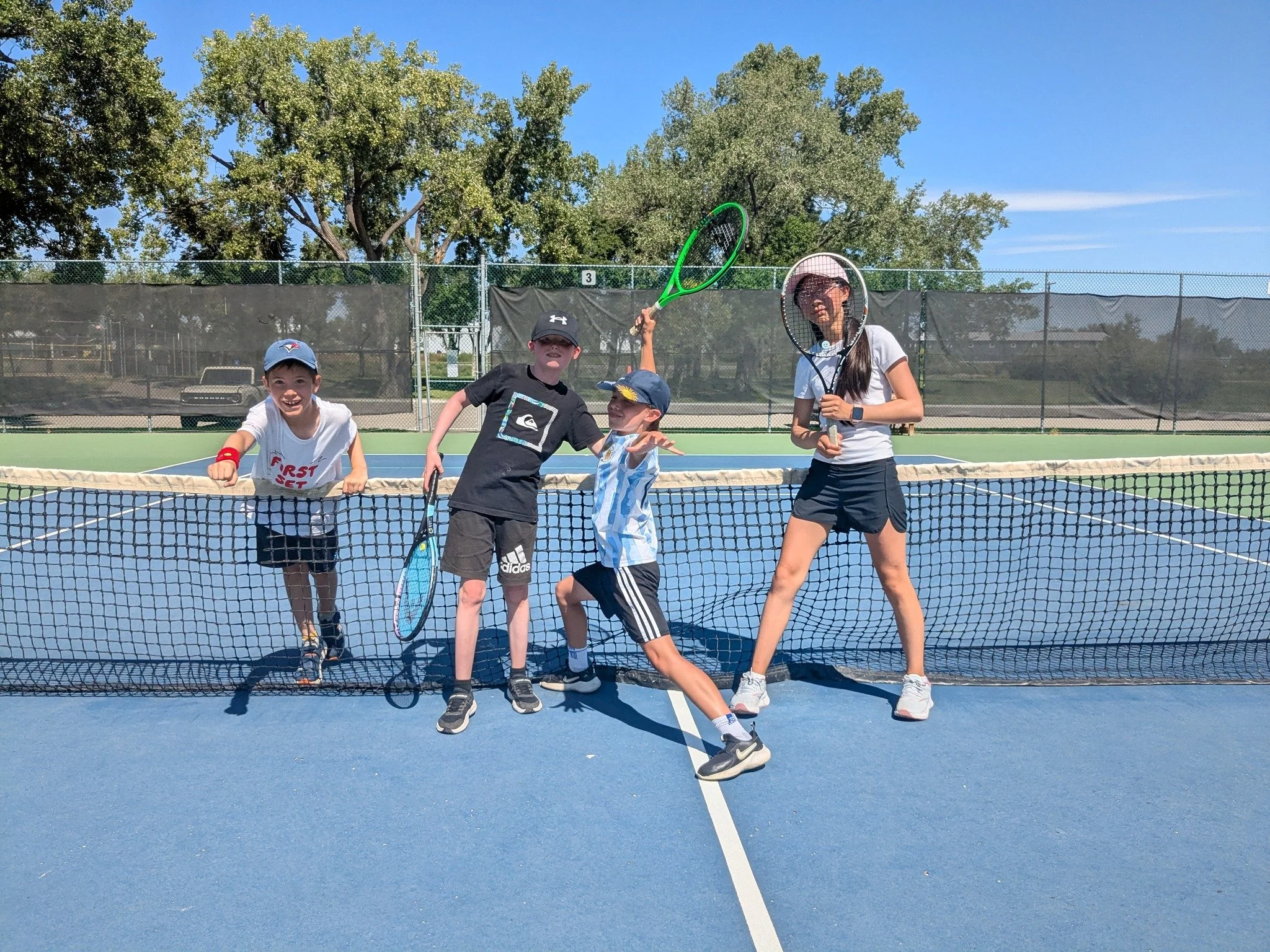Four children on a tennis court, two boys and two girls, playing with court racquets under a sunny sky, with trees in the background.