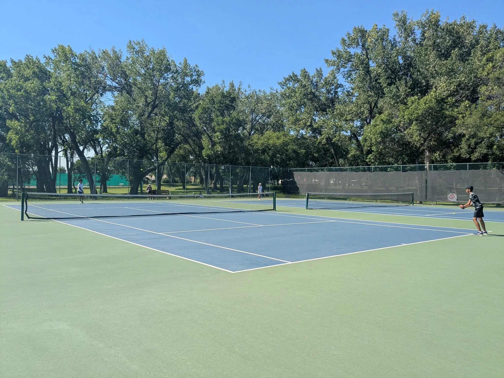 People playing tennis on an outdoor court surrounded by trees and a blue sky.