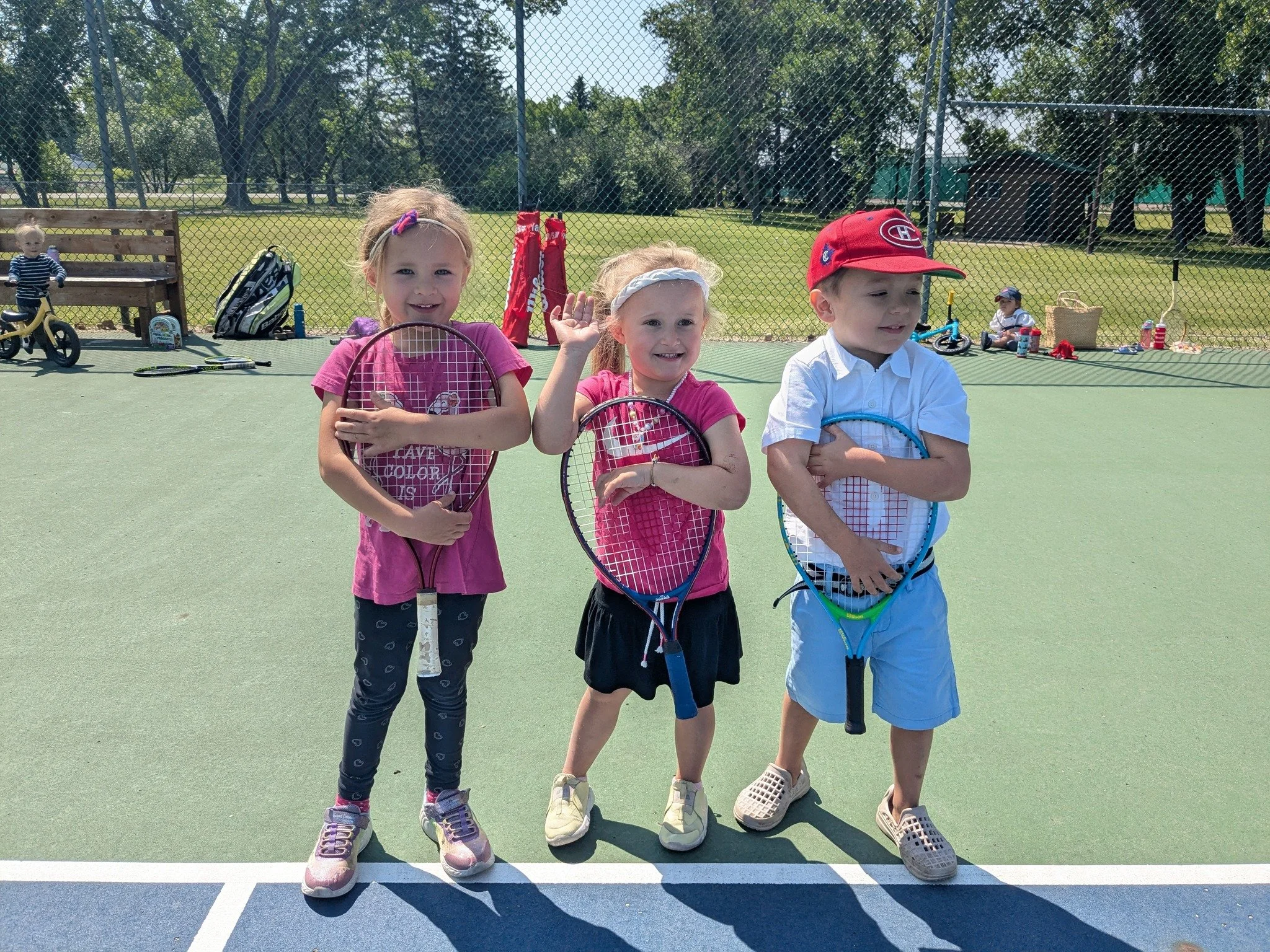 Three children standing on a tennis court holding tennis rackets, with other children playing in the background.