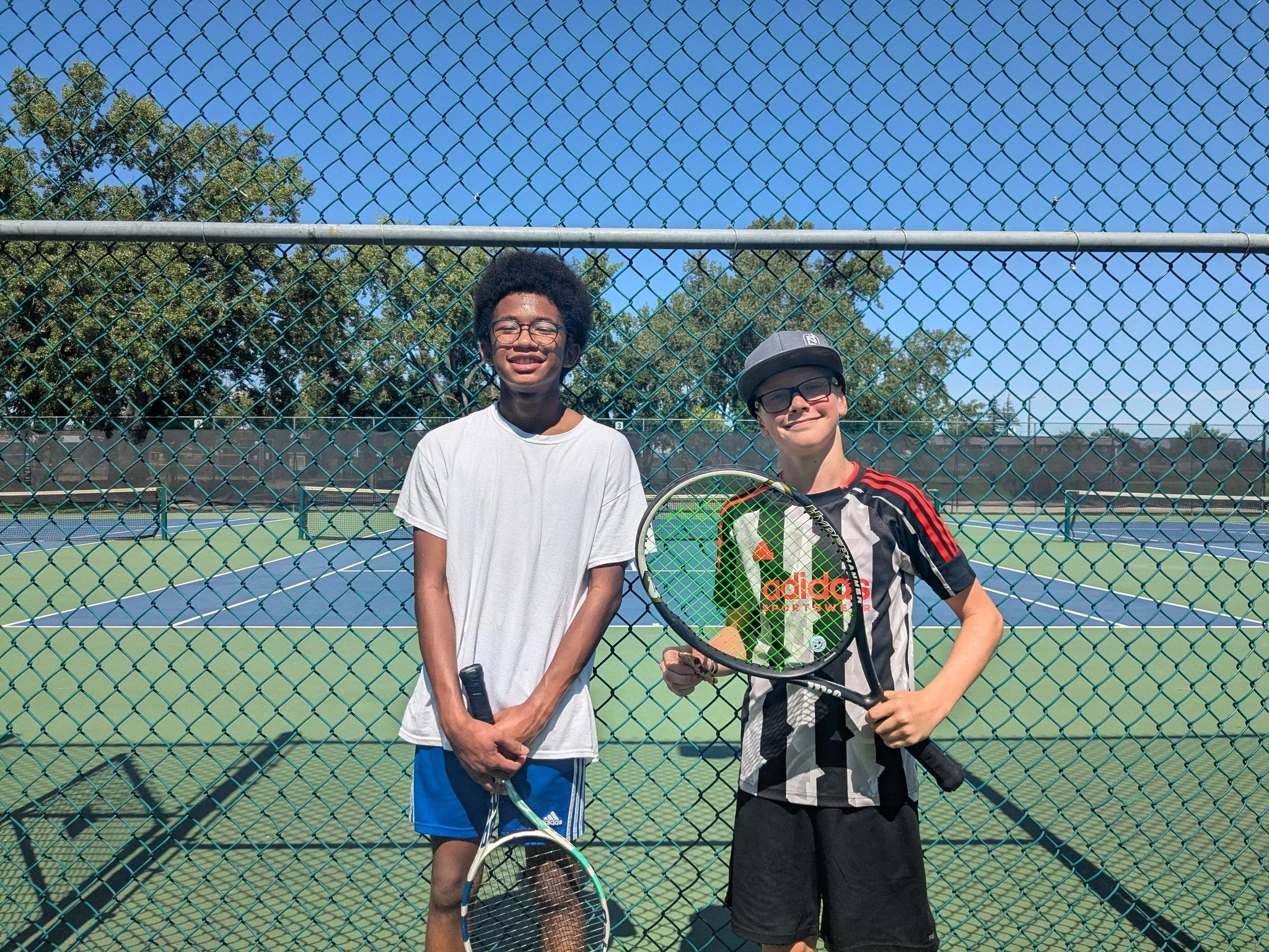 Two young boys standing behind a chain-link fence on a tennis court, holding tennis rackets, smiling, with a clear blue sky and trees in the background.