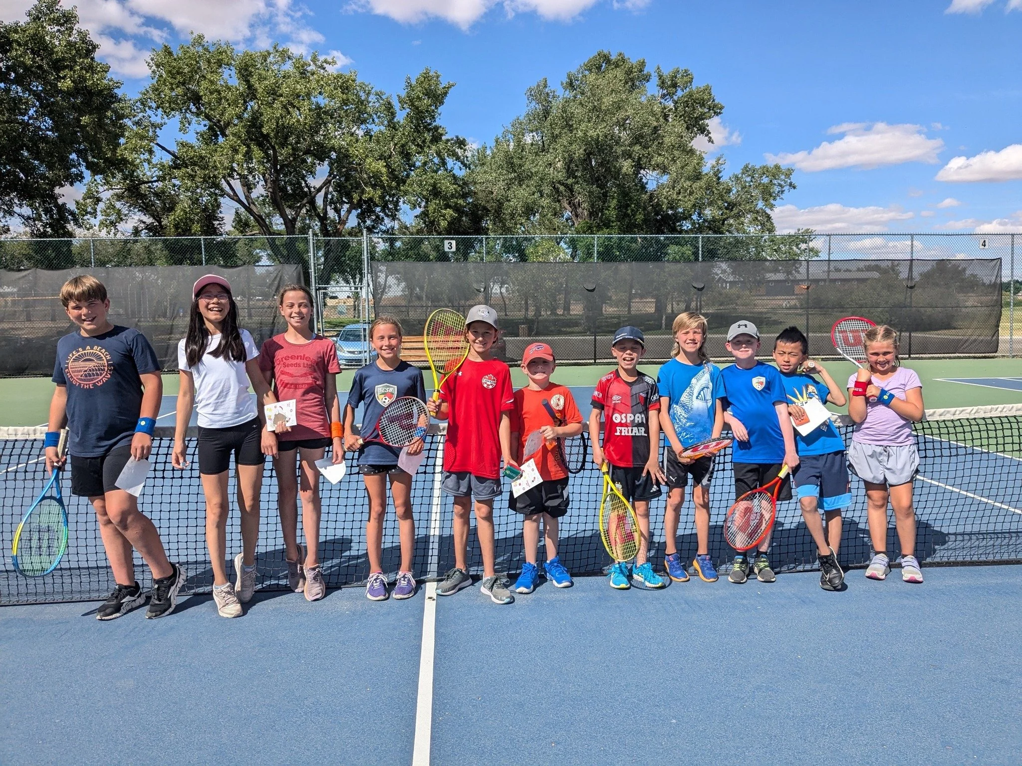 A group of children standing on a tennis court, holding tennis rackets and smiling, with a tennis net in front of them and trees in the background.